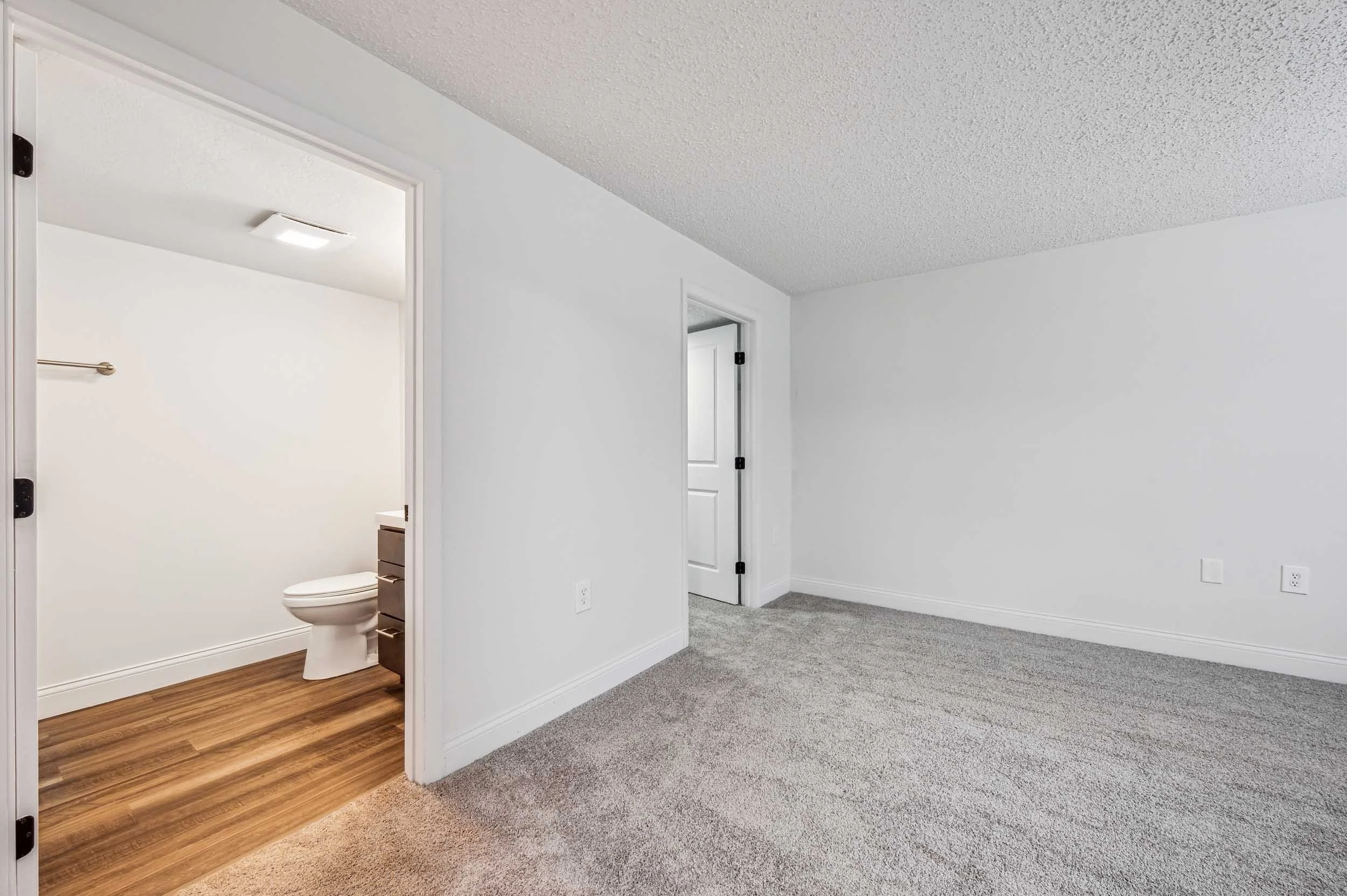Empty room with gray carpet, white walls, and two open doorways, one leading to a bathroom with wooden floor and a toilet visible.