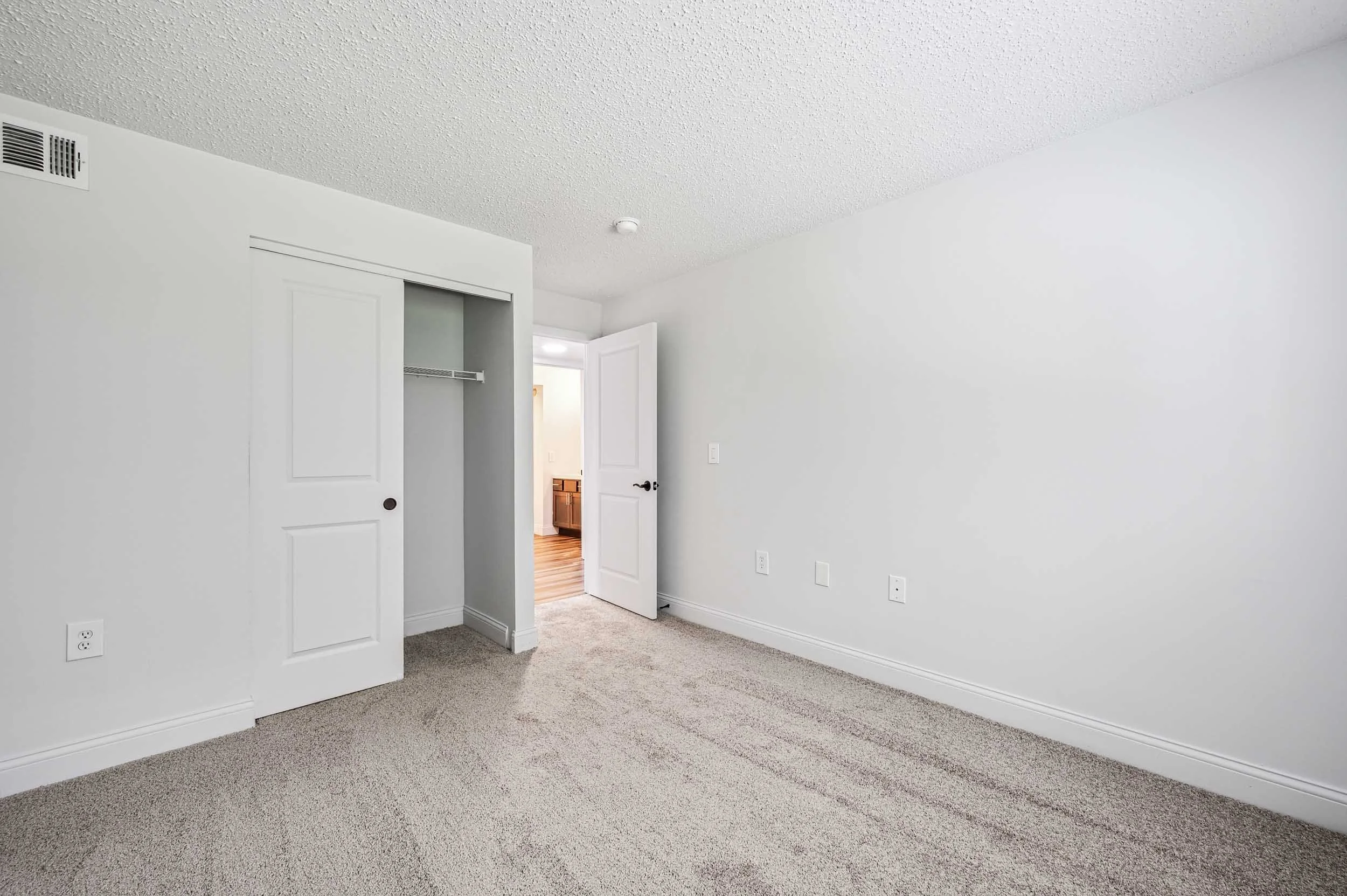 Empty room with beige carpet, white walls, and open closet door, with a view of another room through a doorway.