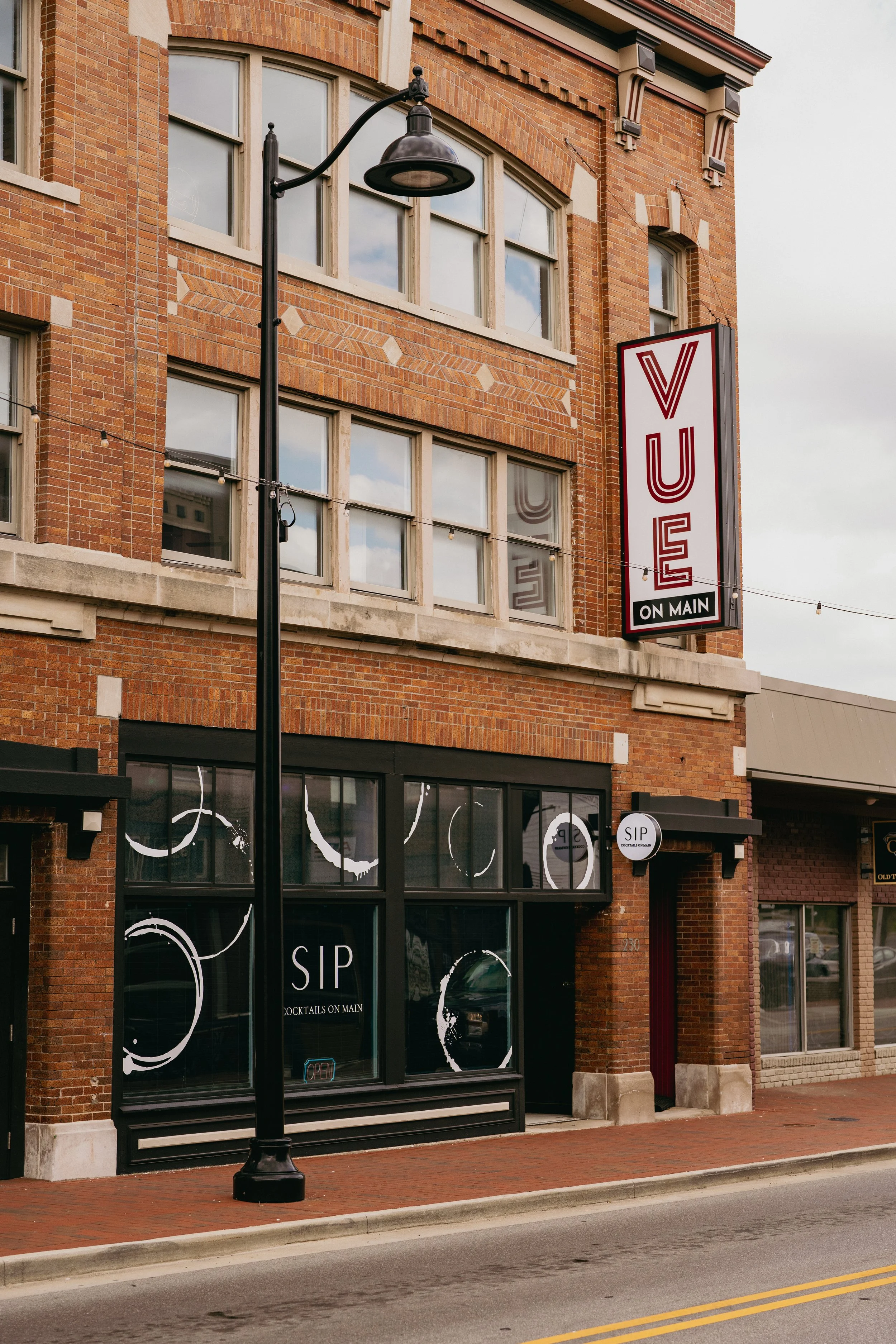 Red brick building with large windows and a sign that reads 'VUE ON MAIN' and 'COCKTAILS ON MAIN.' A black street lamp is in the foreground, and the street has a brick sidewalk and yellow road lines.