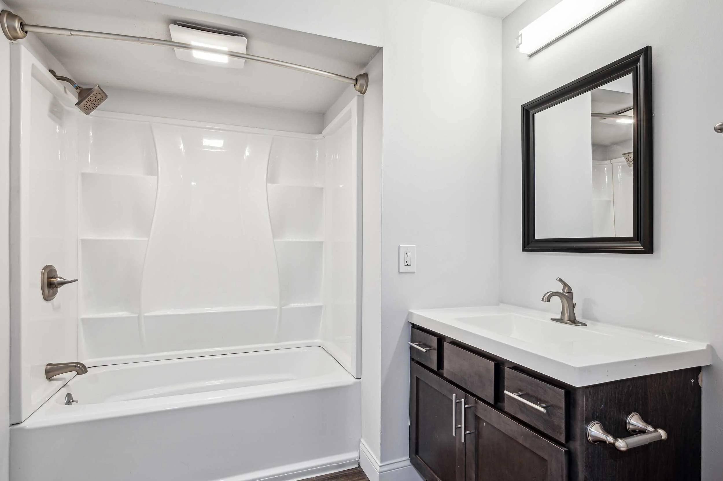 A modern bathroom with a white shower enclosure, a dark wooden vanity cabinet with a white countertop, a rectangular mirror with a black frame, and brass fixtures.