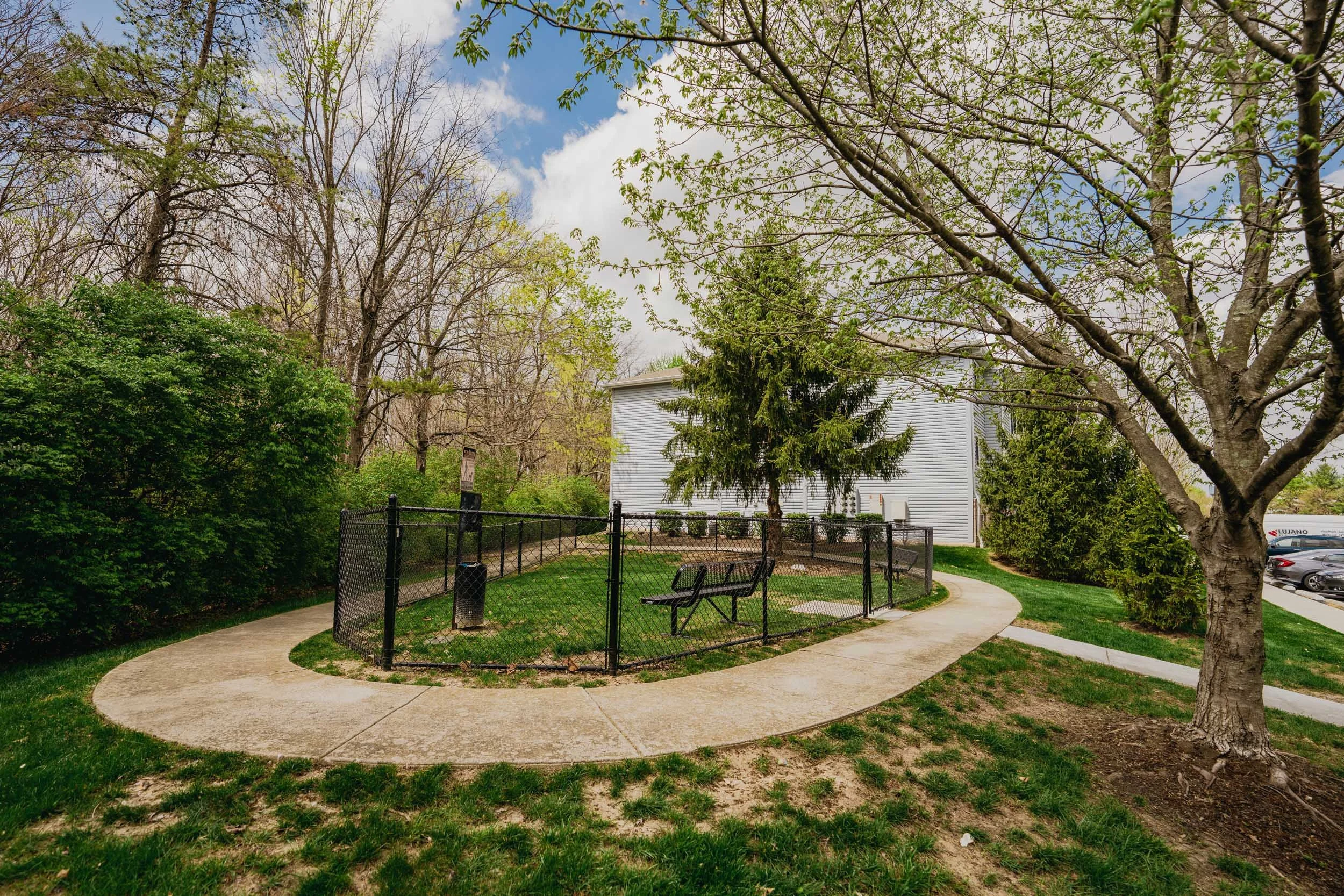 A fenced Dog Park with a concrete path winding through green grass, surrounded by trees and bushes, with a white building and parked cars in the background under a partly cloudy sky.