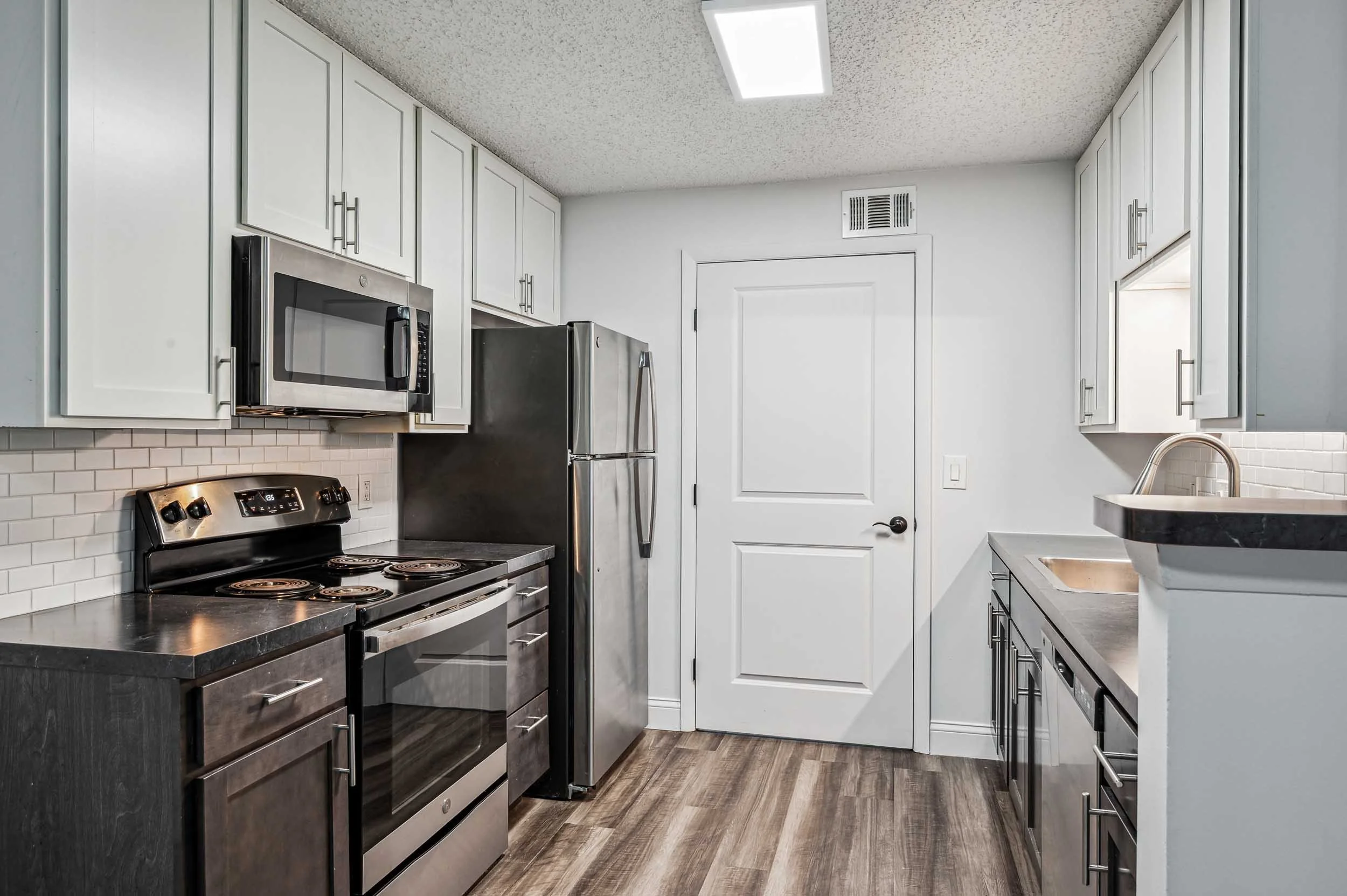 Modern kitchen with white upper cabinets, dark lower cabinets, stainless steel appliances including a microwave, refrigerator, and stove, white subway tile backsplash, and wood-look laminate flooring.