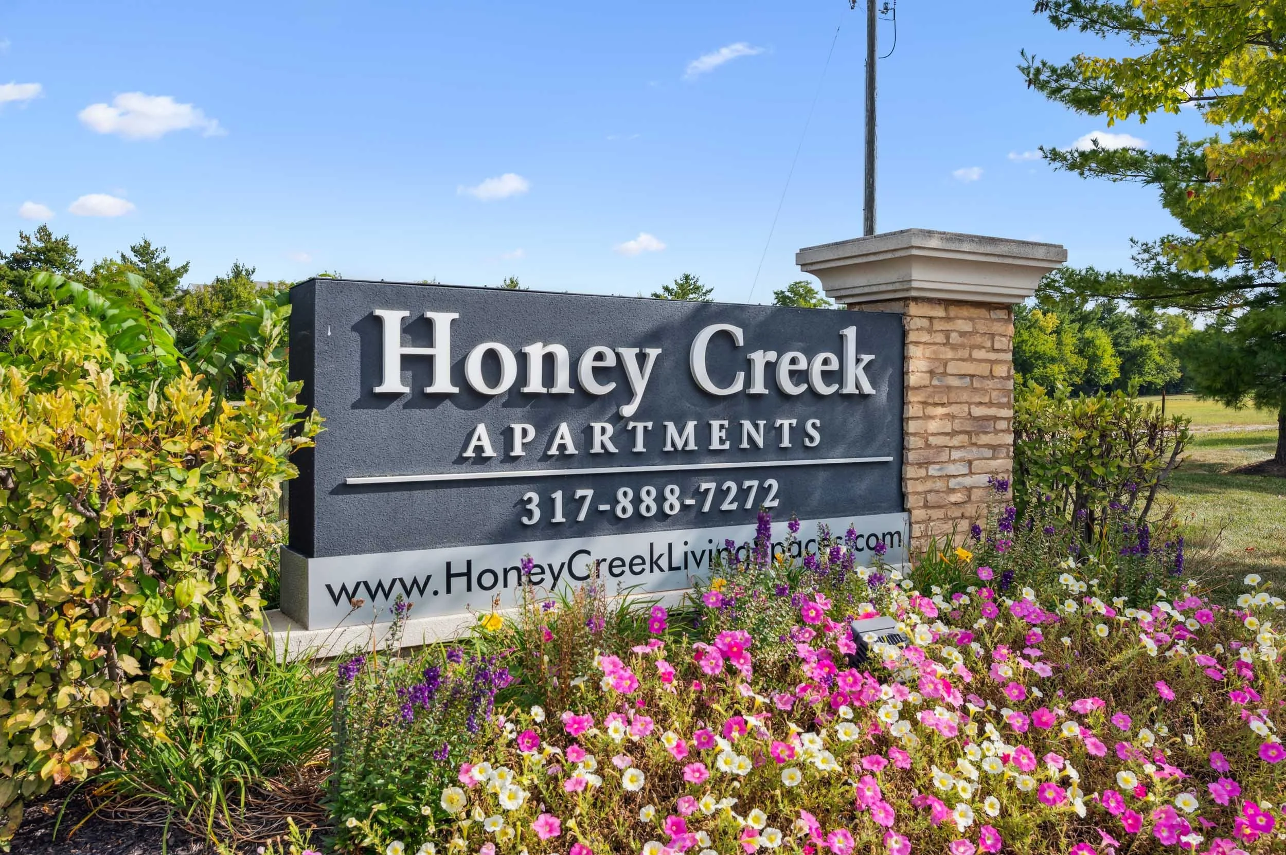 Sign for Honey Creek Apartments surrounded by colorful flowers and greenery on a sunny day.