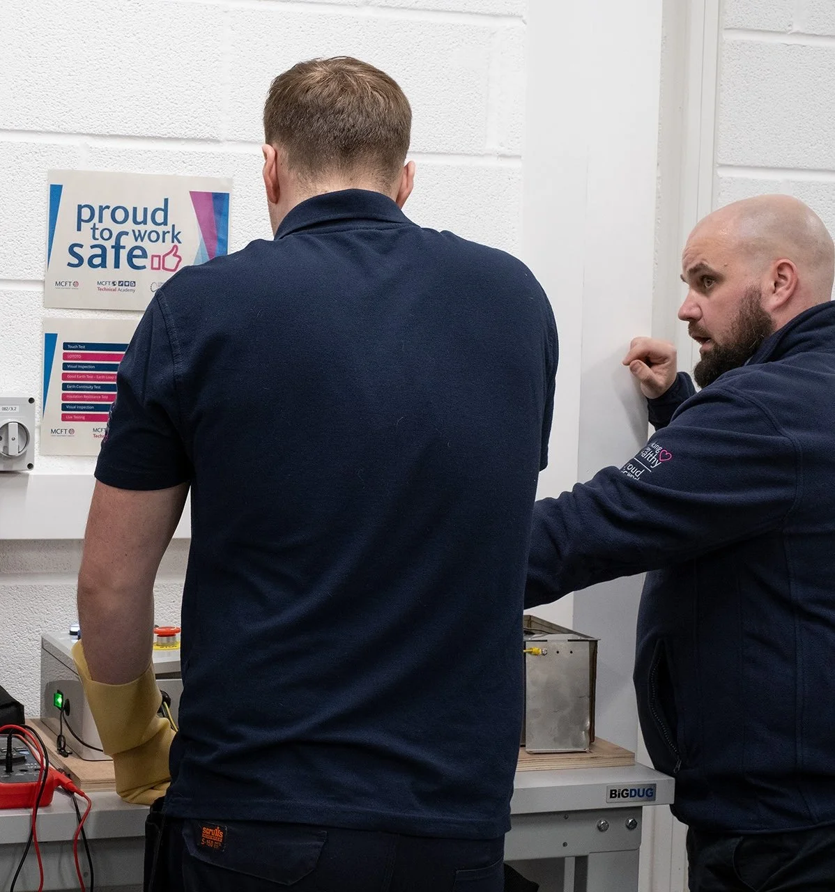Two men working on electrical equipment in a lab or workshop. One man is facing away, wearing a navy blue shirt with a yellow glove on his left arm. The other man, with a bald head and beard, is facing sideways and points towards the equipment.