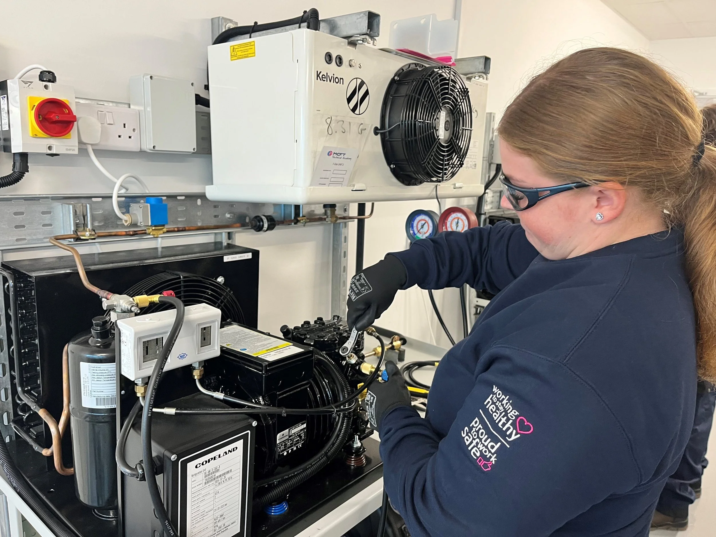A woman working on HVAC equipment, wearing safety glasses and gloves, with industrial tools and gauges around her.