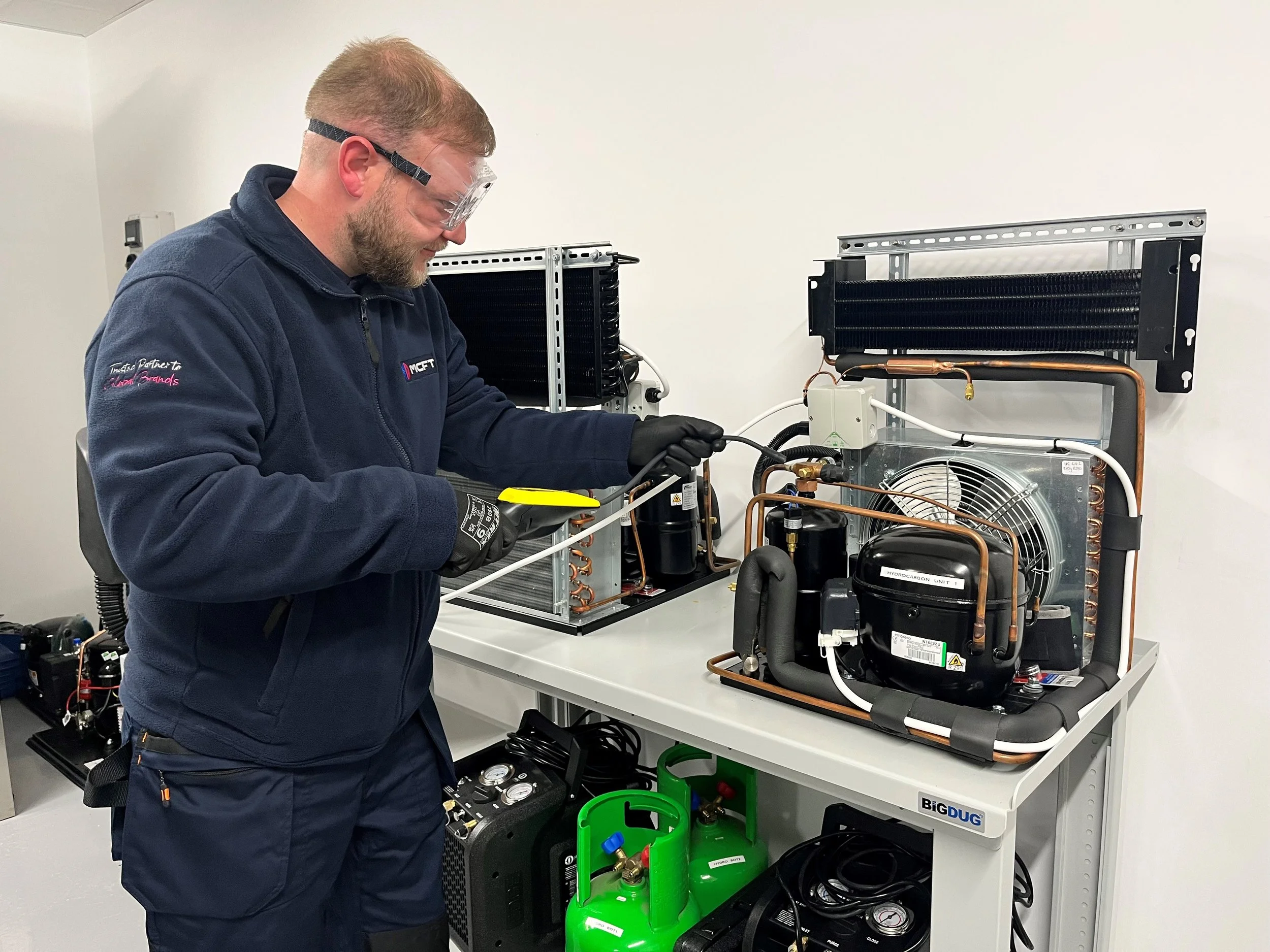 A technician working on an HVAC or refrigeration system, using tools and wearing safety glasses, in a clean workshop.