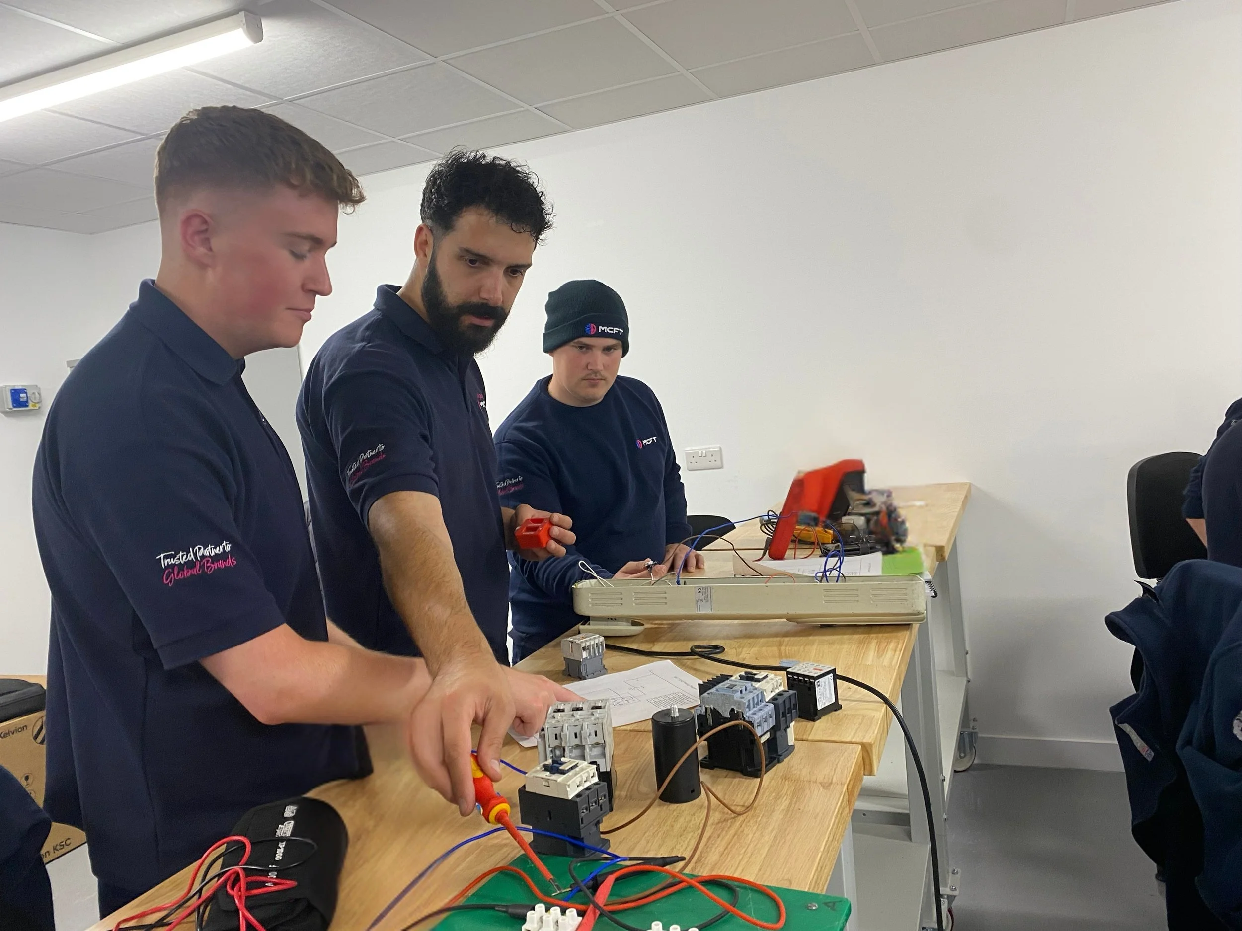 Three young men in navy blue shirts working on electrical wiring and components at a wooden table, in an indoor classroom or training room setting.