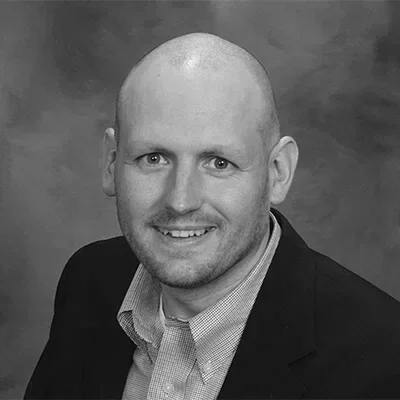 Black and white portrait of a smiling man with a shaved head, wearing a suit jacket and collared shirt.
