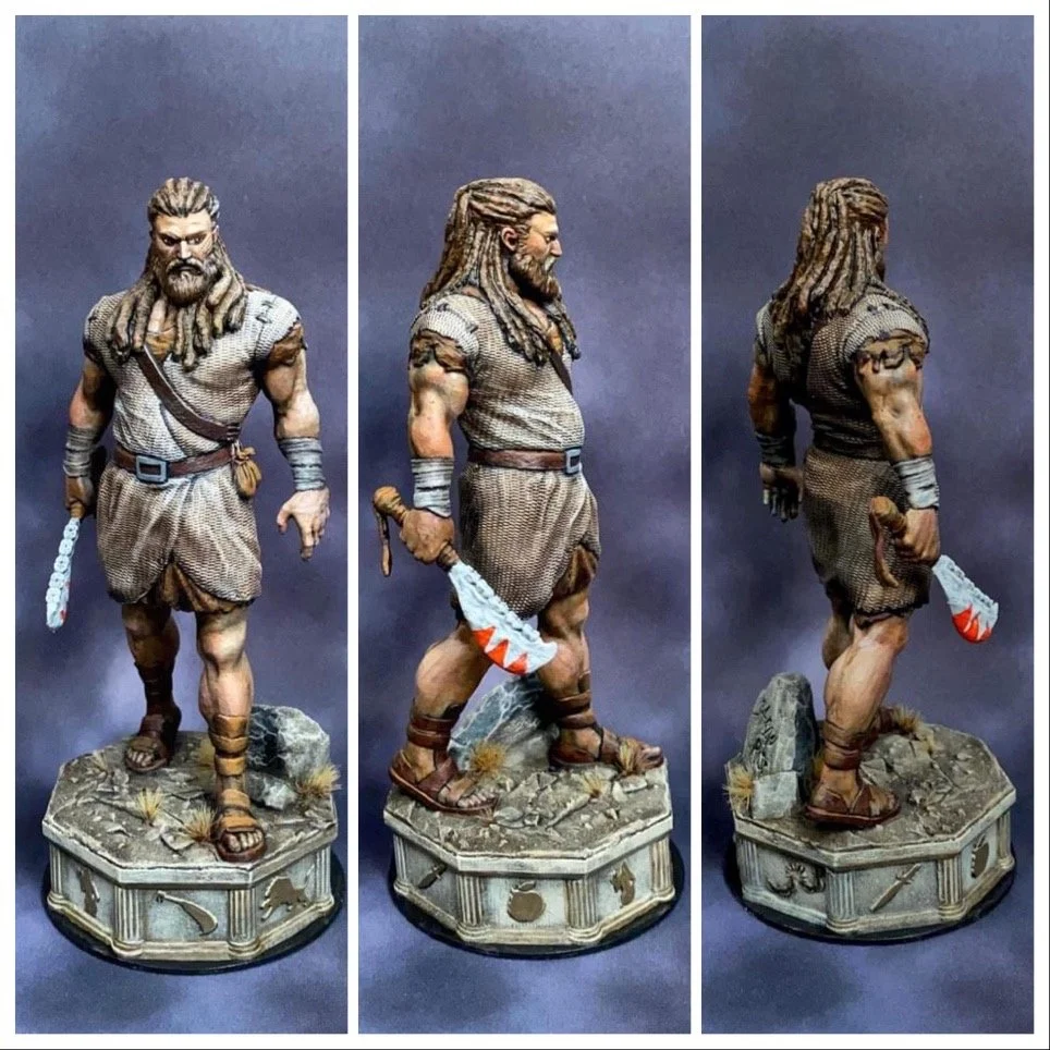 Detailed figurine of a muscular warrior with long dreadlocks, beard, wearing tribal-style clothing, sandals, and holding a blood-stained weapon, standing on a decorated base with rocks and grass.