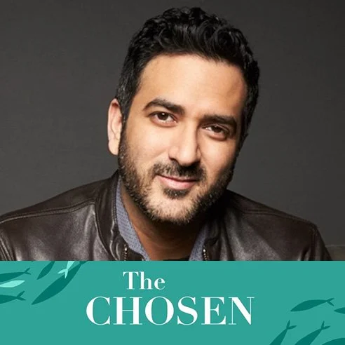 A man with dark hair and facial hair, wearing a leather jacket and a collared shirt, smiling at the camera with a plain dark background. Text overlay says 'The Chosen'.