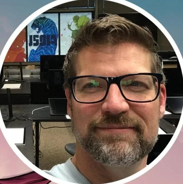 A man with glasses and a beard smiling in a computer lab with colorful dinosaur artwork on the window in the background.