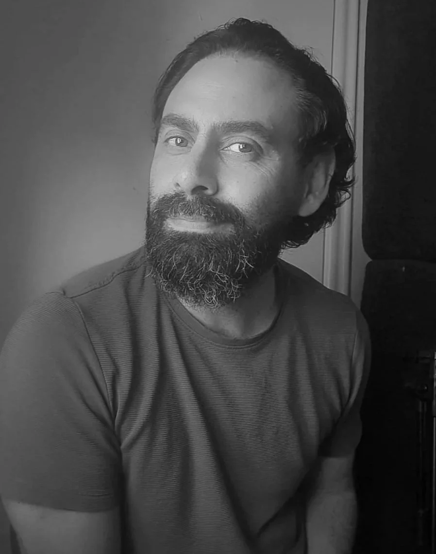 Black and white portrait of a man with a beard and mustache, looking towards the camera with a slight smile, wearing a textured t-shirt, sitting in front of a plain wall and a doorframe.