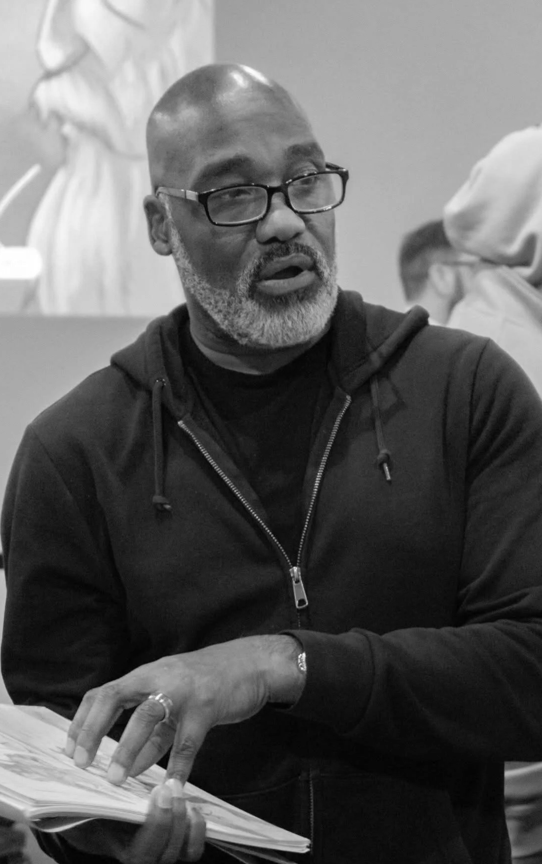 A bald African American man with glasses and a gray beard, wearing a black hoodie, appears to be talking and gesturing with a book in front of him.