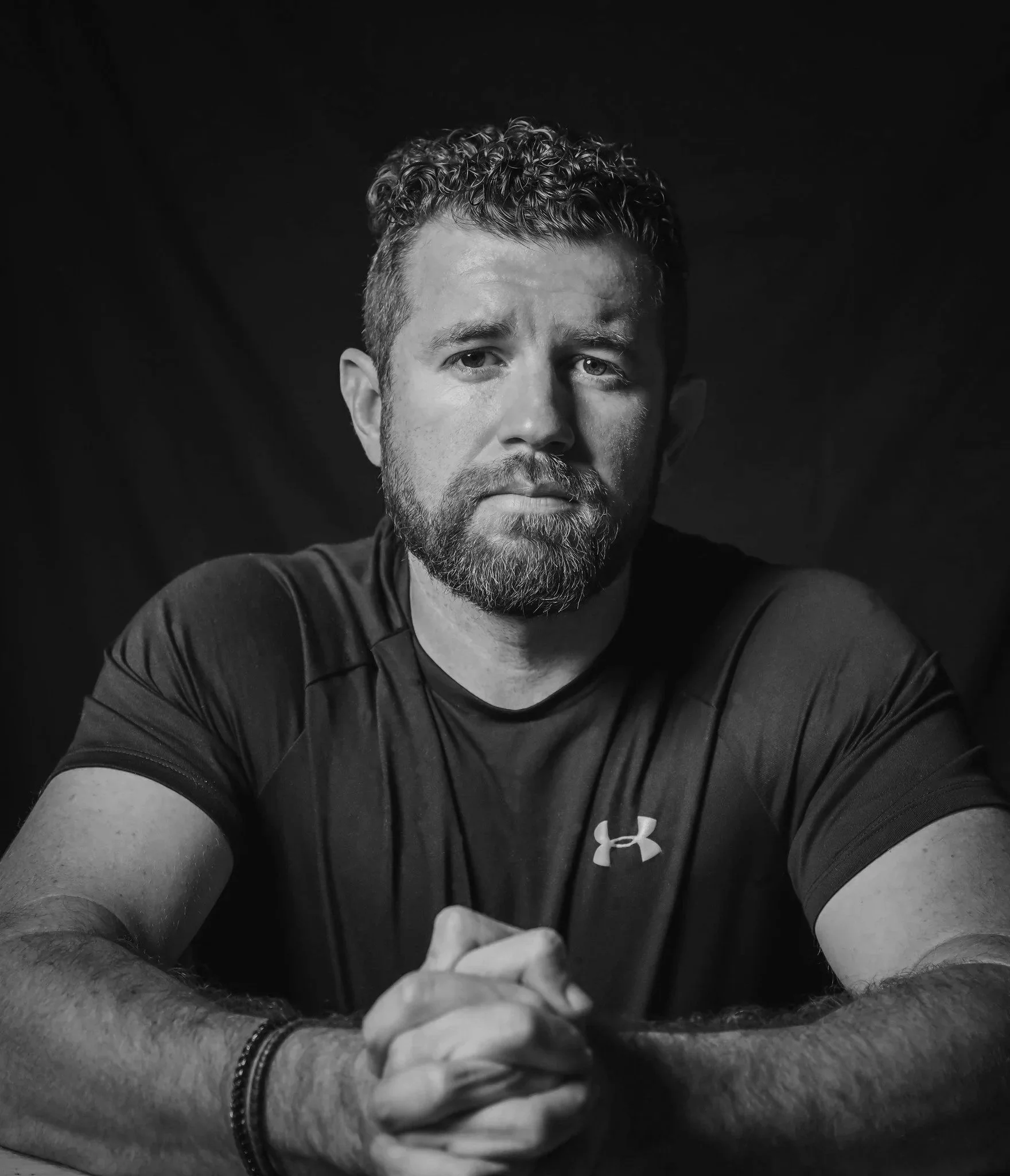 A man with a beard and short curly hair taking a selfie, wearing a dark shirt and a bracelet, with a serious expression against a dark background.