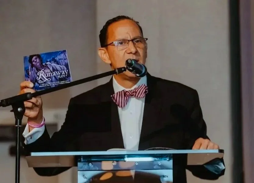 A man in a tuxedo and red bow tie speaking at a podium, holding a book titled "The Runaway Plot".