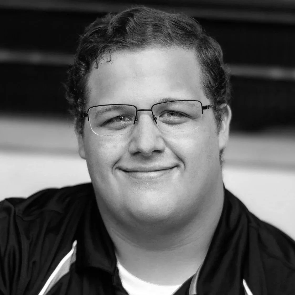 A black and white portrait of a smiling man with glasses and short wavy hair.