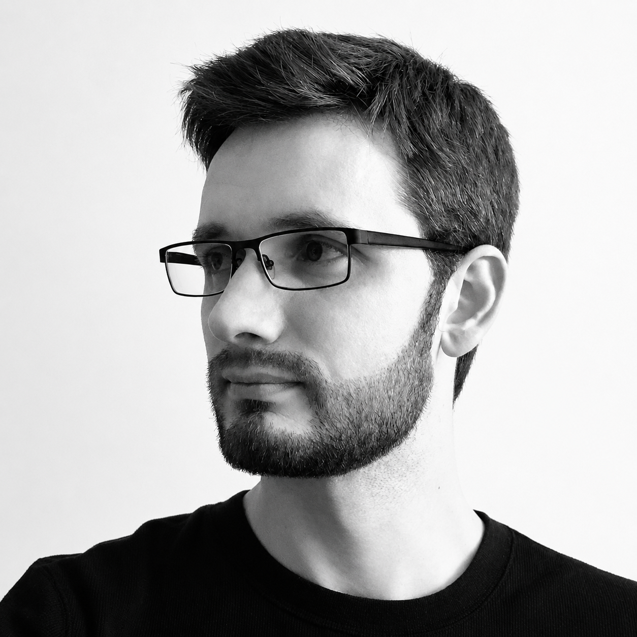 Black and white portrait of a young man with short dark hair, glasses, and a beard, wearing a black shirt, looking to the left against a plain white background.