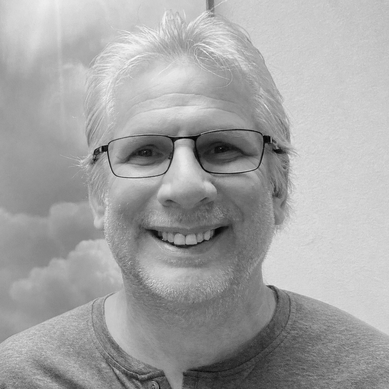 Black and white photo of a smiling man with glasses, short gray hair, and a beard, wearing a casual shirt, standing in front of a backdrop with clouds.