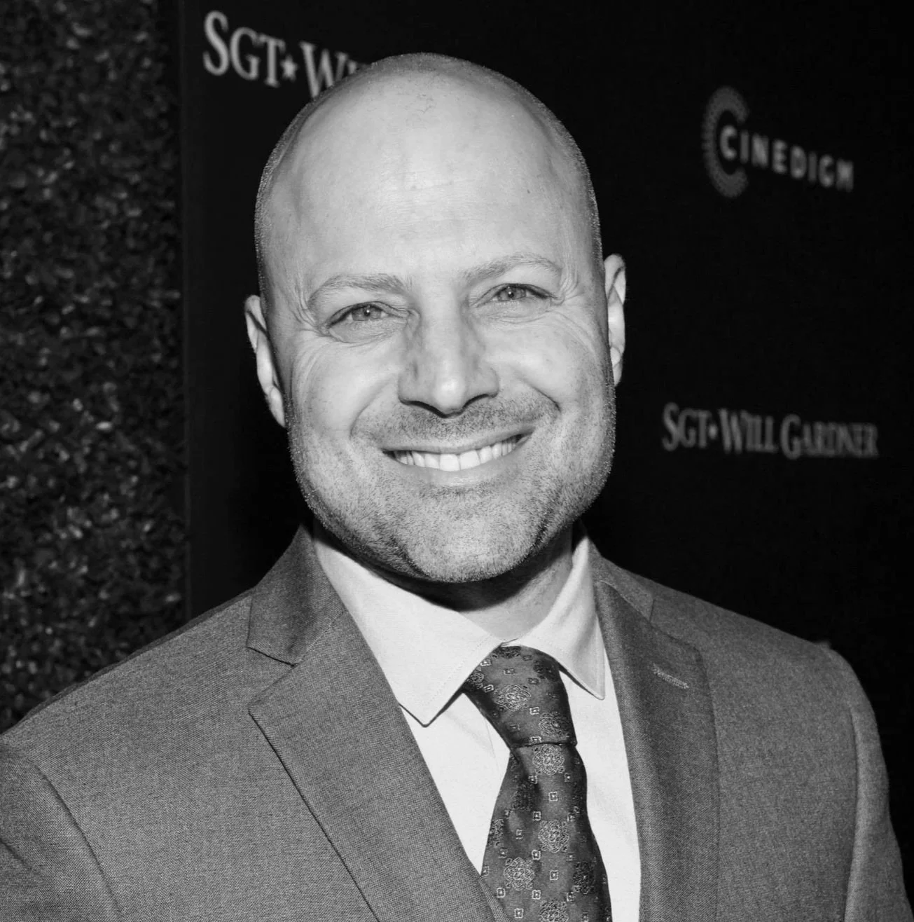 Black and white photo of a smiling man in a suit and tie, standing in front of a backdrop with text.