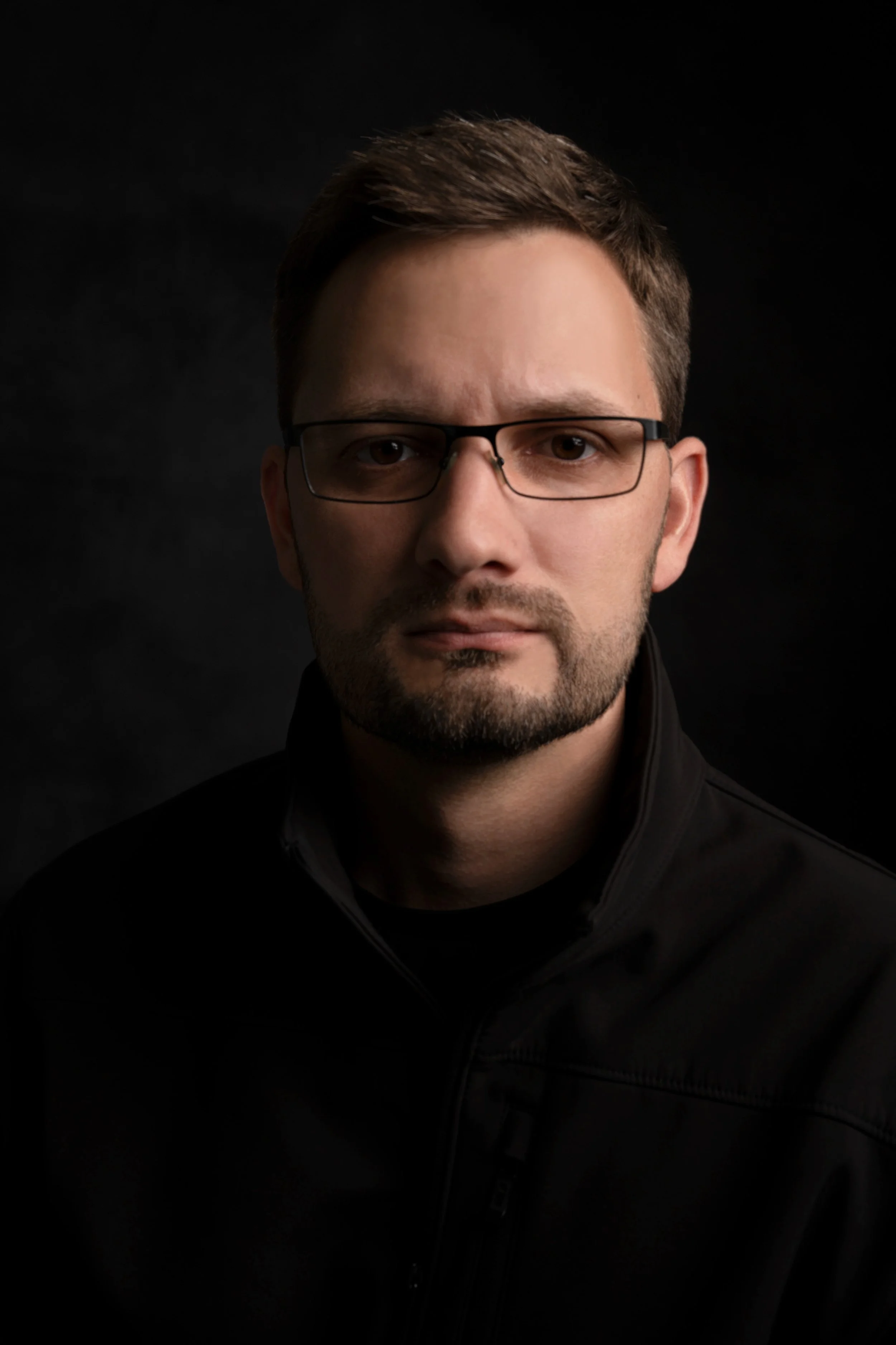 Close-up portrait of a young man with brown hair, glasses, and a beard, wearing a black jacket, against a dark background.