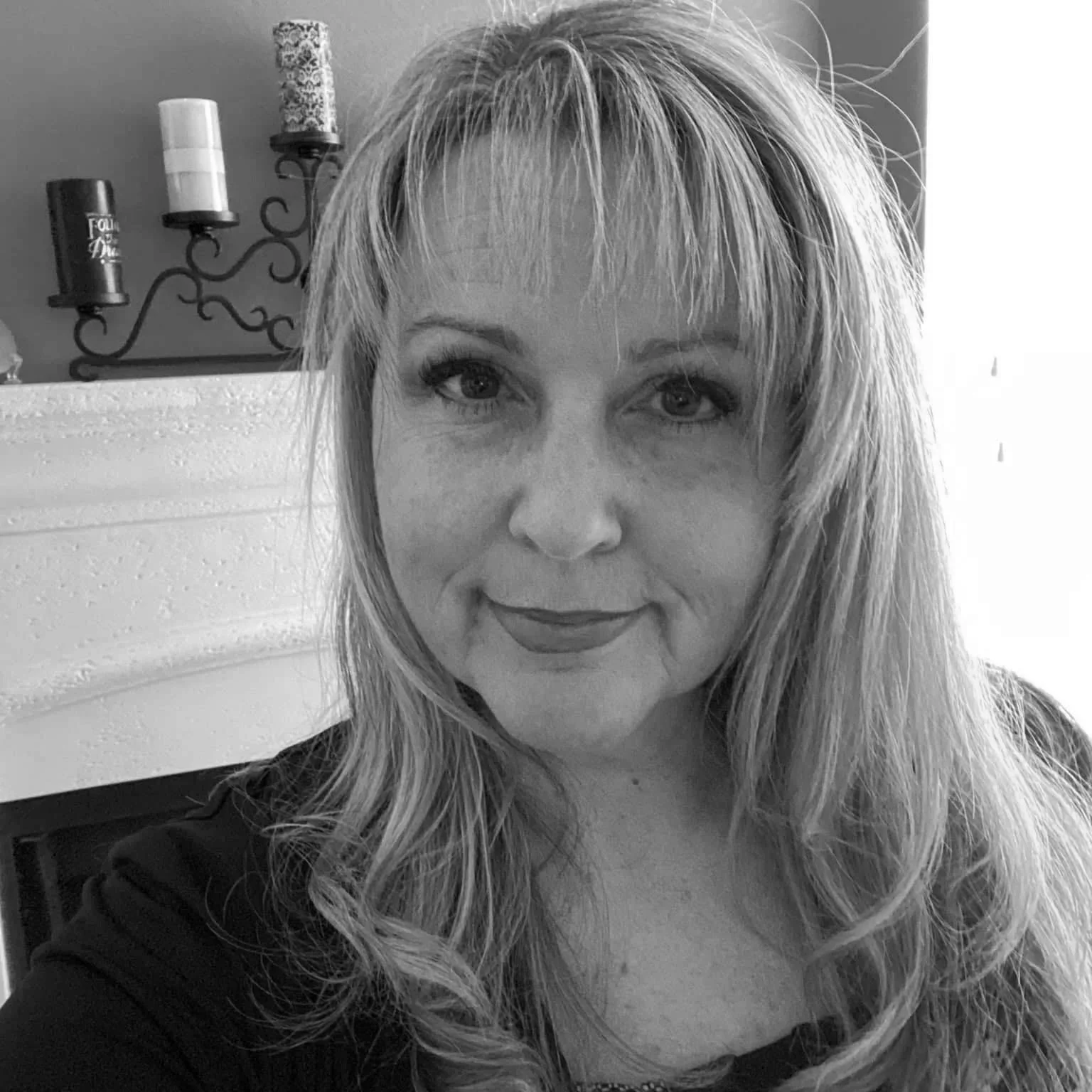 A black and white photo of a woman with wavy hair, smiling slightly, indoors near a wall with candle holders.