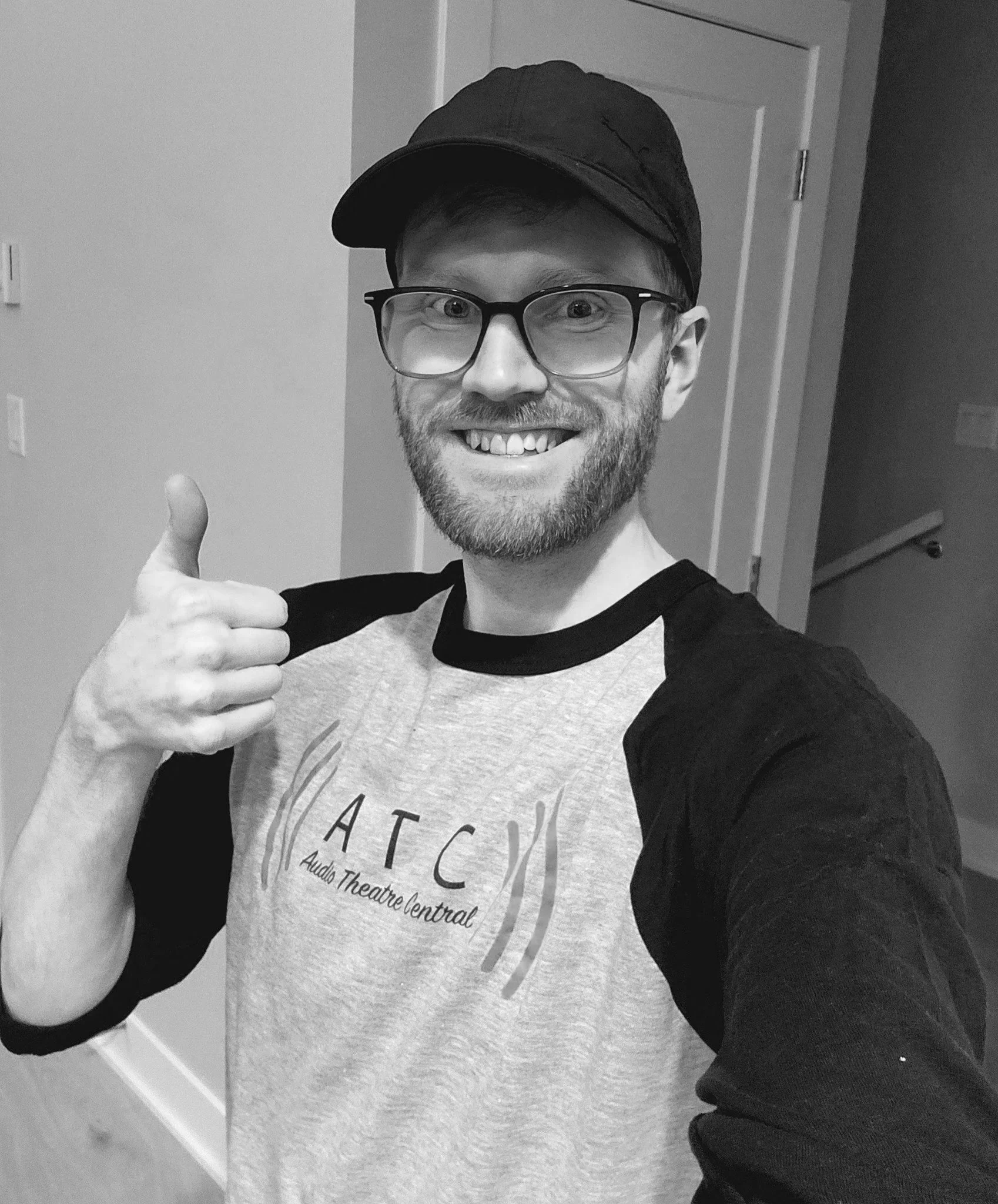 A man with a beard and glasses smiling and giving a thumbs-up gesture in a room with plain walls, wearing a black baseball cap and a baseball-style shirt.