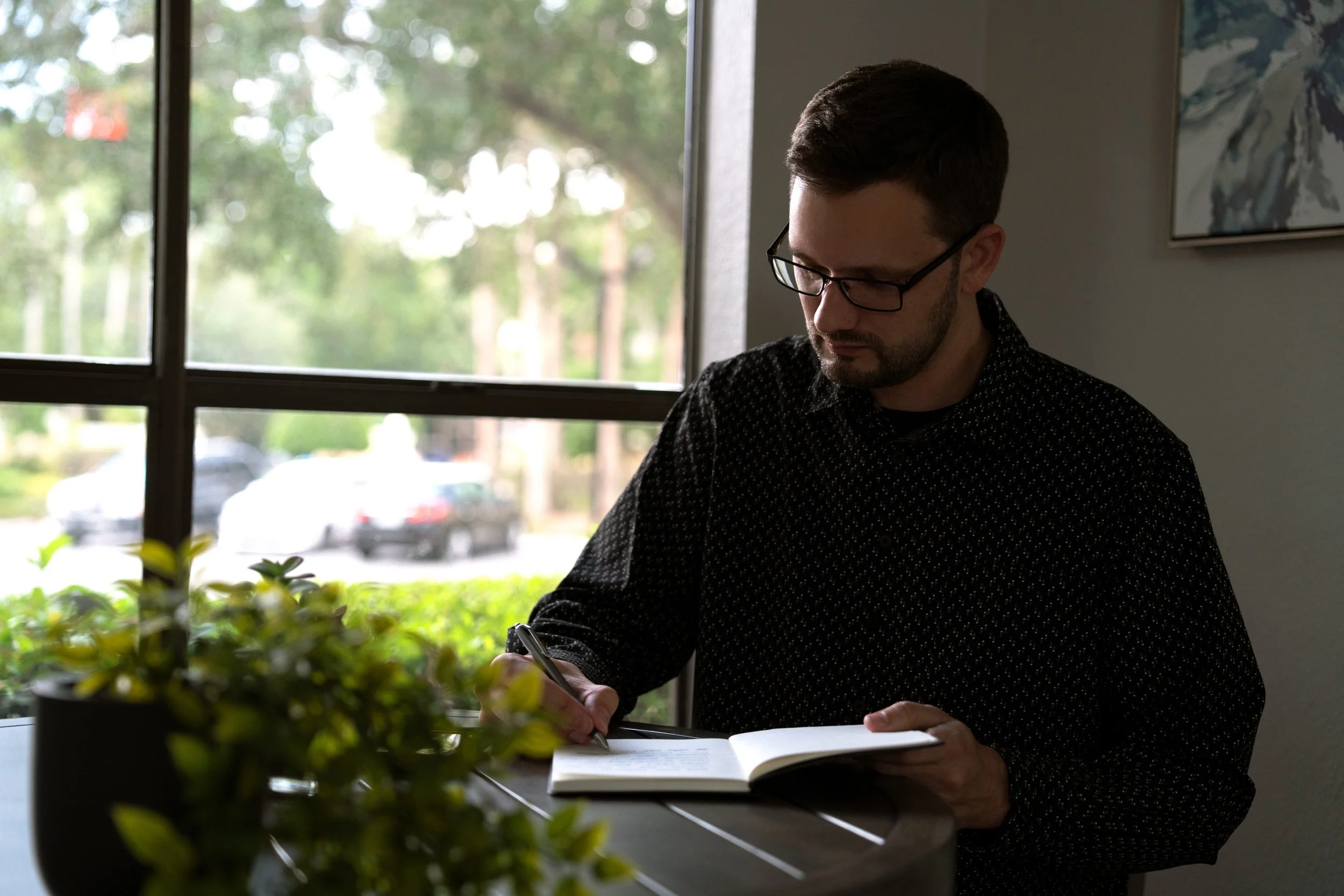 A man with dark hair, glasses, and a beard sitting at a table near a window, reading or writing in a book with a potted plant in the foreground.