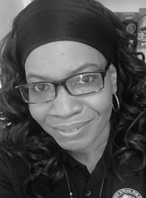 A woman with curly hair wearing glasses, a headband, hoop earrings, and a uniform with a badge, smiling in a close-up black and white photo.