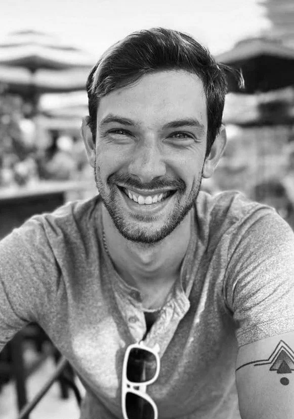 A smiling young man with short hair and a beard, wearing a T-shirt and sunglasses hanging from his shirt, sitting outdoors at a cafe with umbrellas in the background.