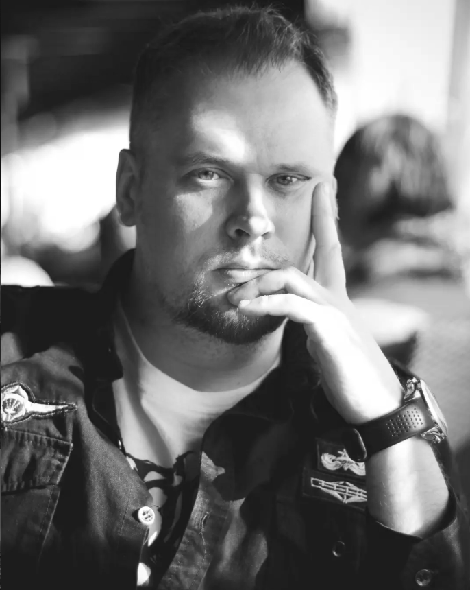 Black and white photo of a man with short hair and a beard resting his face on his hand, wearing a dark jacket with patches and a watch, sitting indoors.