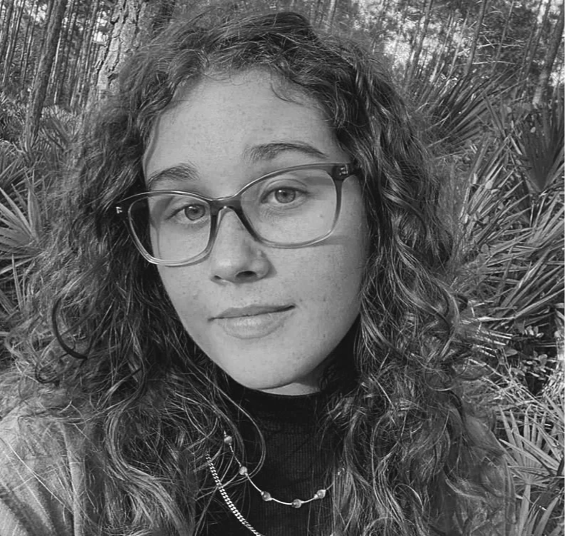 A young woman with curly hair and glasses, standing outdoors with a background of dense trees and plants.