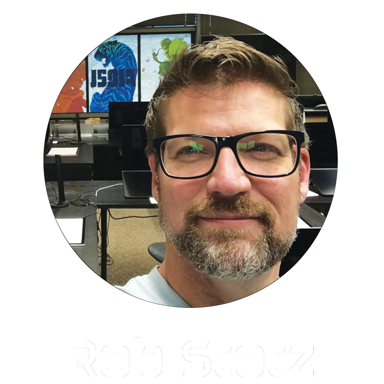 A man with glasses, a beard, and short brown hair taking a selfie in a classroom environment with desktop computers and colorful artwork in the background, with the name 'Rob Stotz' written below.