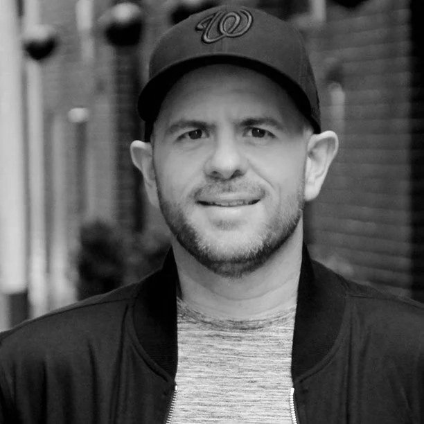 A man wearing a baseball cap with a 'W' logo, a dark jacket, and a light shirt, smiling outdoors in an urban setting.
