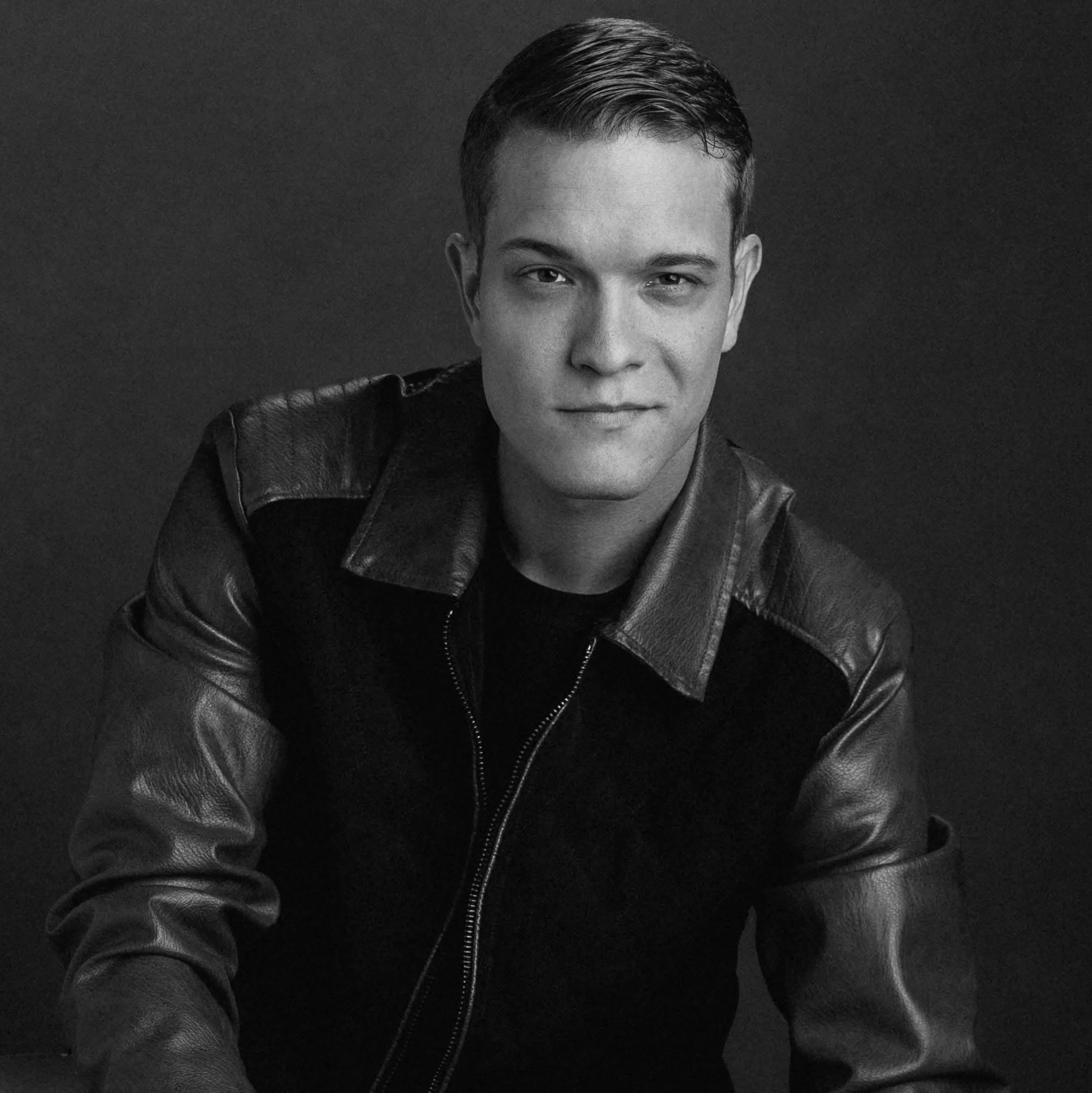 A young man in a black leather jacket sitting on a black leather chair, looking directly at the camera with a confident expression, against a plain dark background.