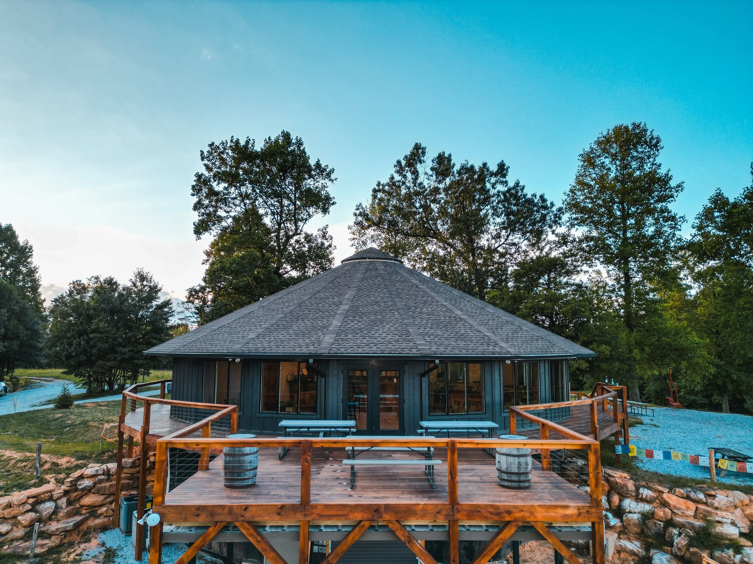 A round, dark-colored building with large windows and a shingled, conical roof, situated on a wooden deck with picnic tables and barrel planters, surrounded by green trees and a gravel area.
