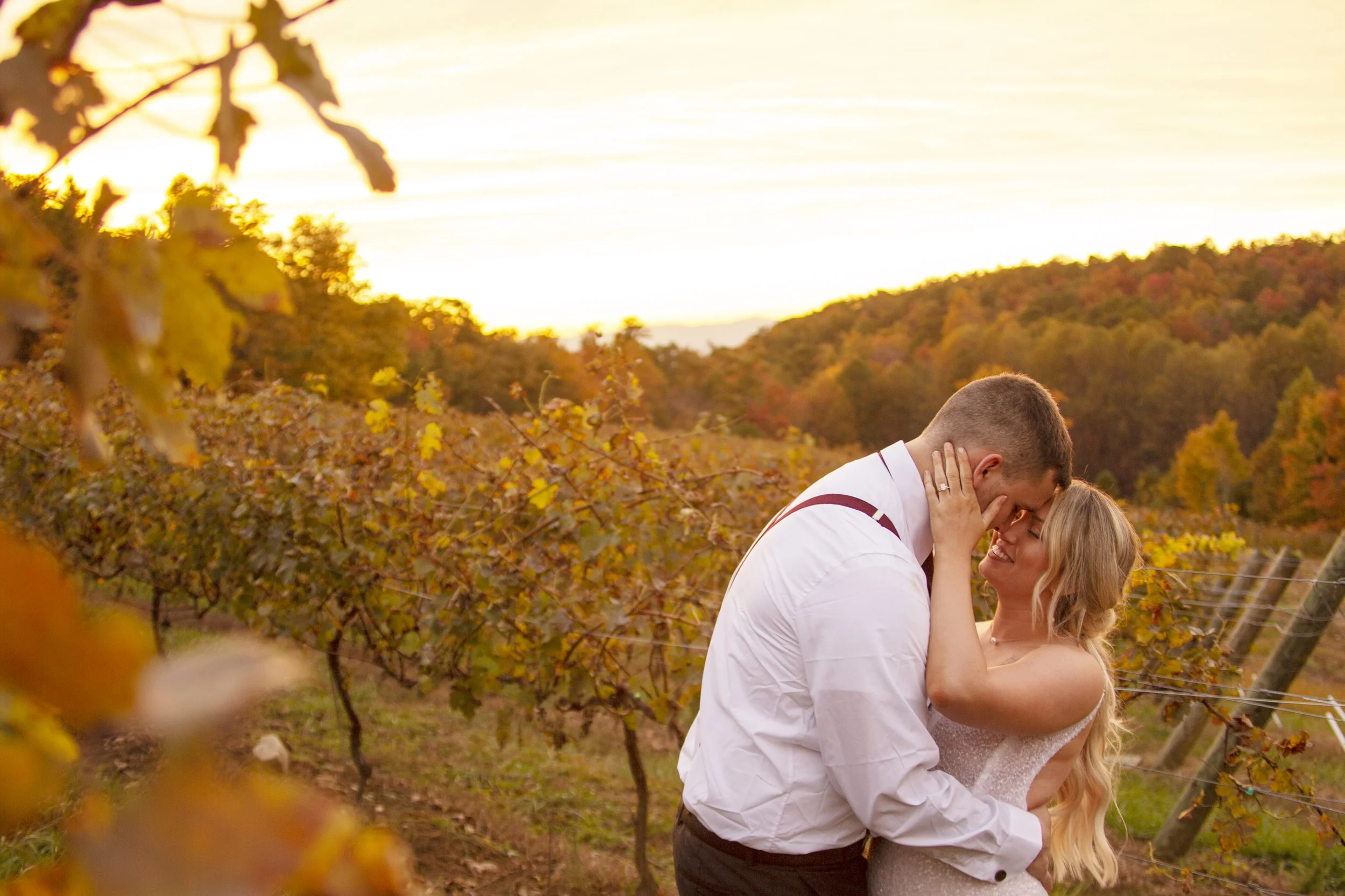 A couple sharing a romantic moment in a vineyard during sunset, with the man holding the woman's face and both smiling, surrounded by autumn foliage.