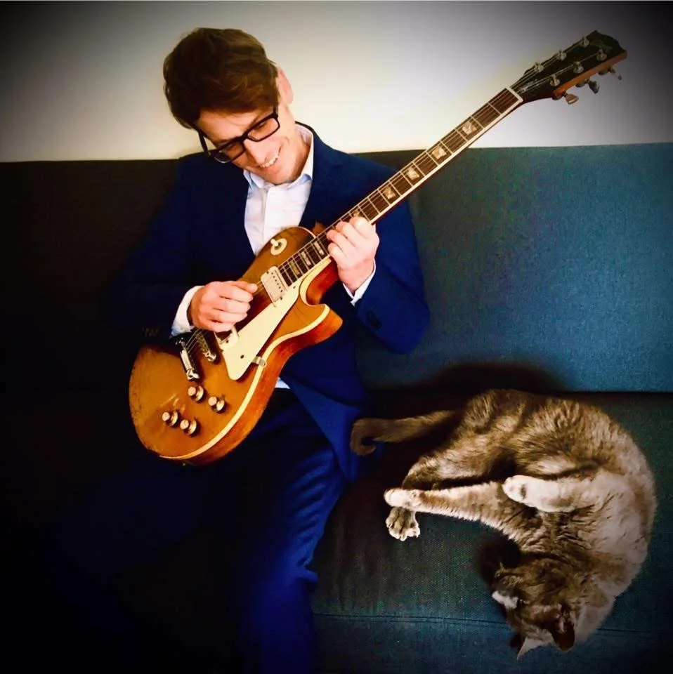 A man standing outdoors in front of an orange vintage Volkswagen van, playing an acoustic guitar.