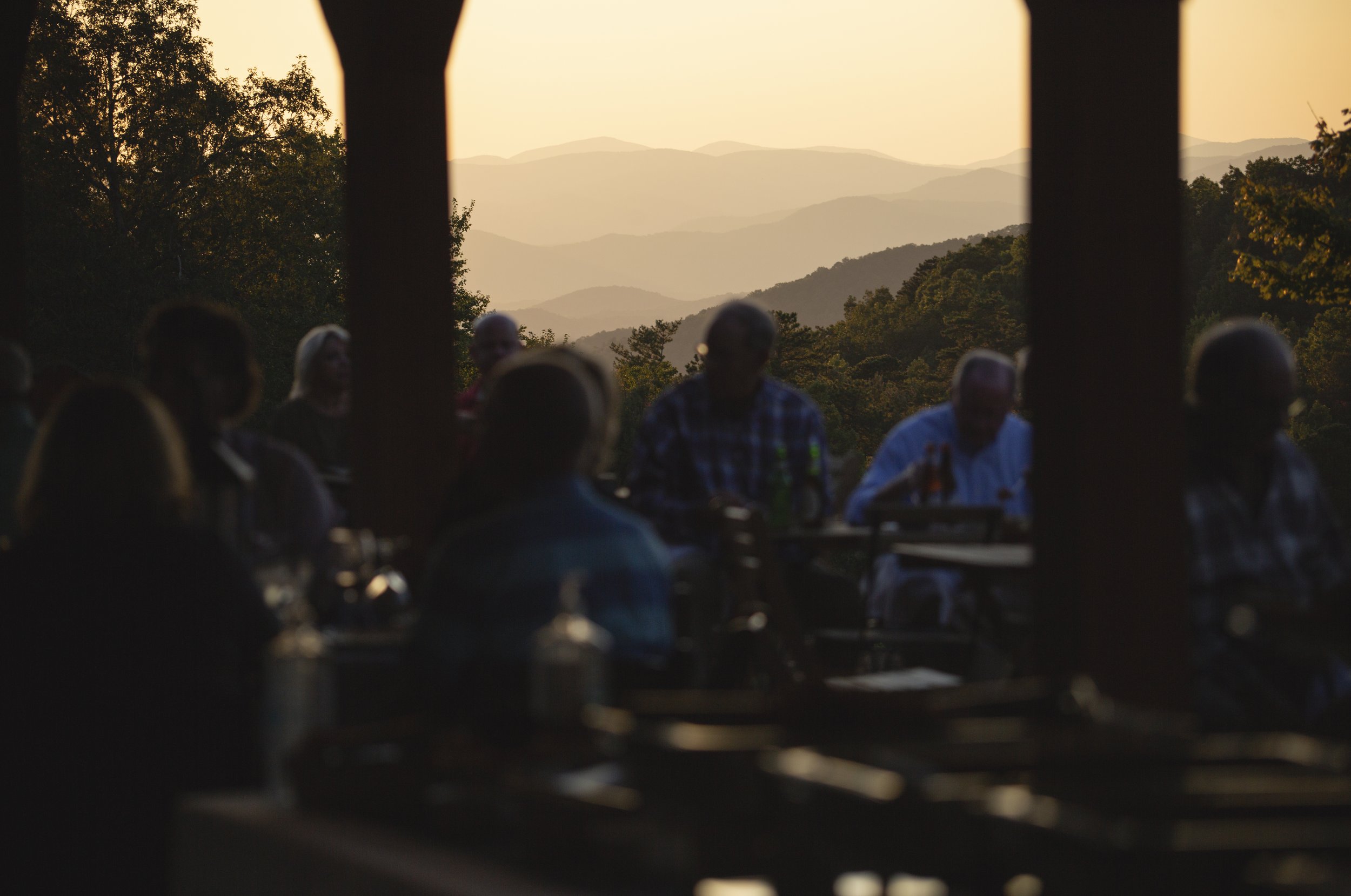 People gathered at an outdoor location during sunset, with mountain ranges and trees in the background.