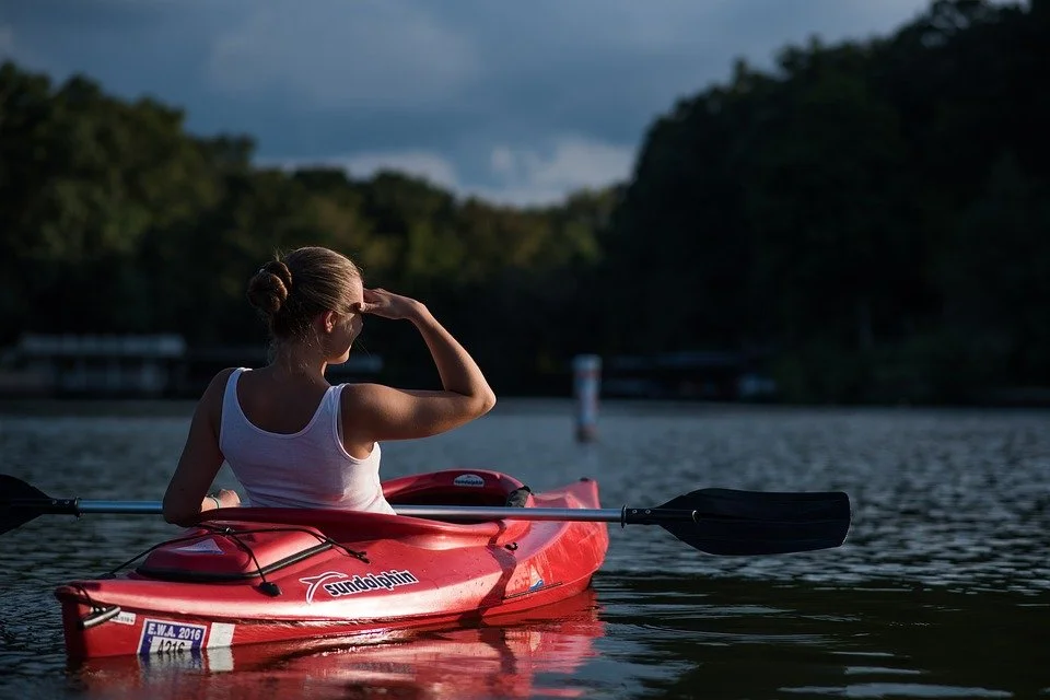 A woman sitting in a red kayak on a calm body of water, looking toward the horizon with her hand shielding her eyes from the sun, surrounded by lush green trees and a cloudy sky.