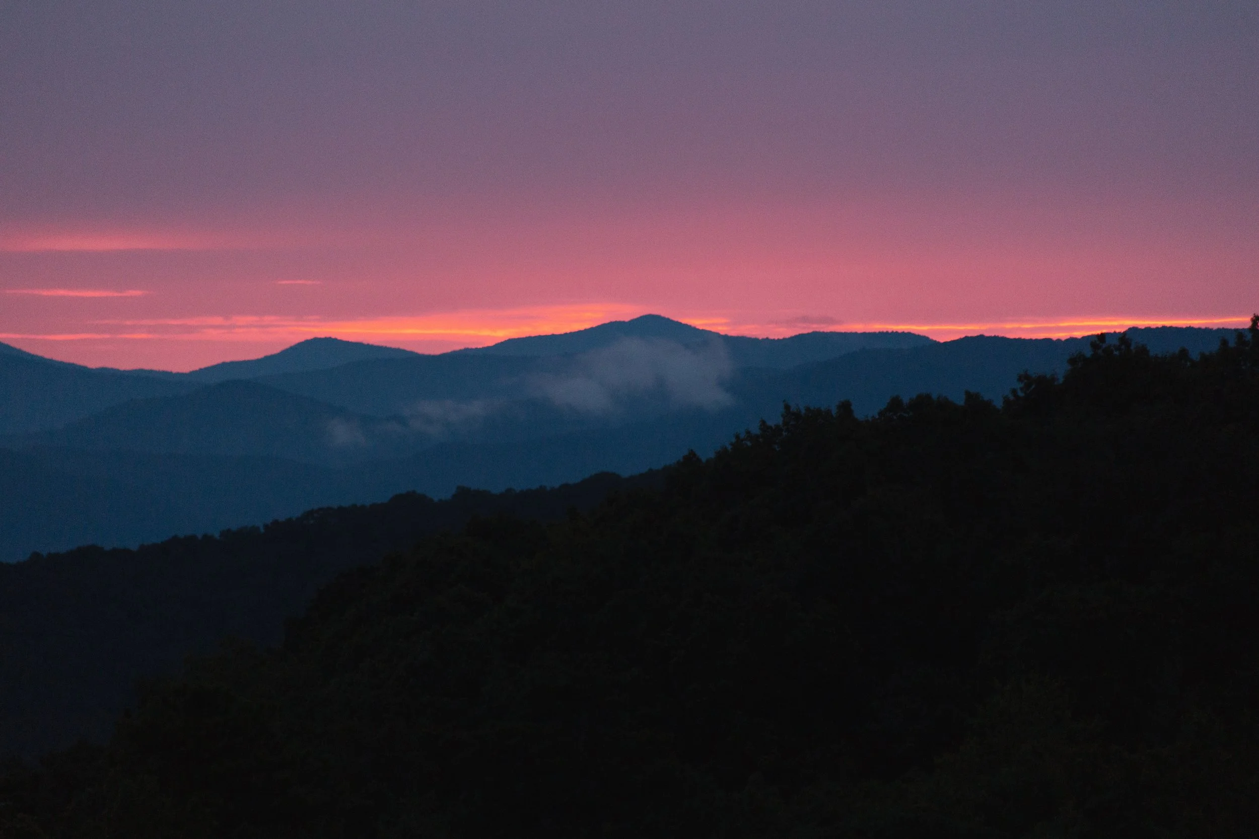 A mountain landscape during sunset with a pink and purple sky, dark silhouettes of mountains, and a misty valley in the foreground.