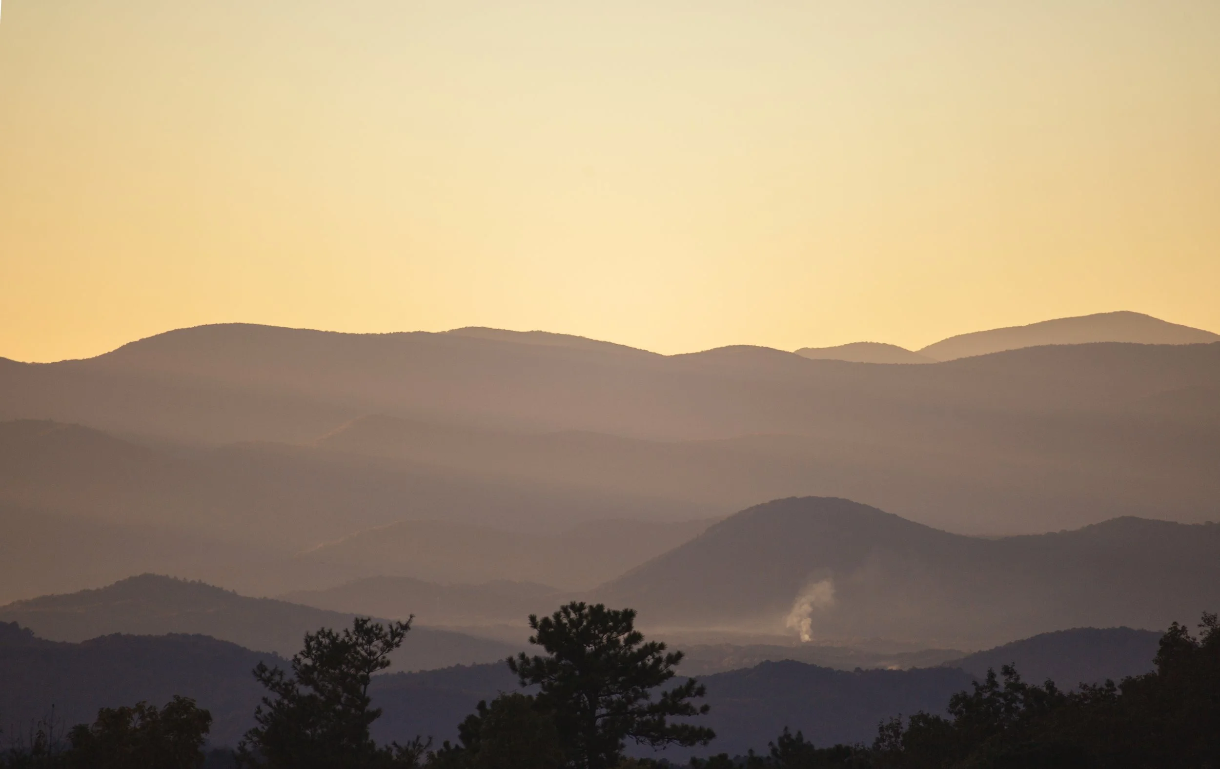 Hazy mountain range at sunrise with trees in the foreground and smoke rising from a distant chimney.