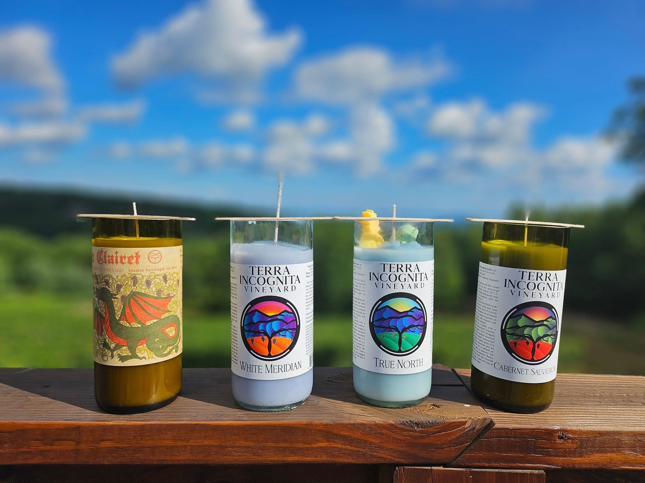 Four colorful candles with wine labels, placed on a wooden surface outdoors with a blue sky and green landscape in the background.
