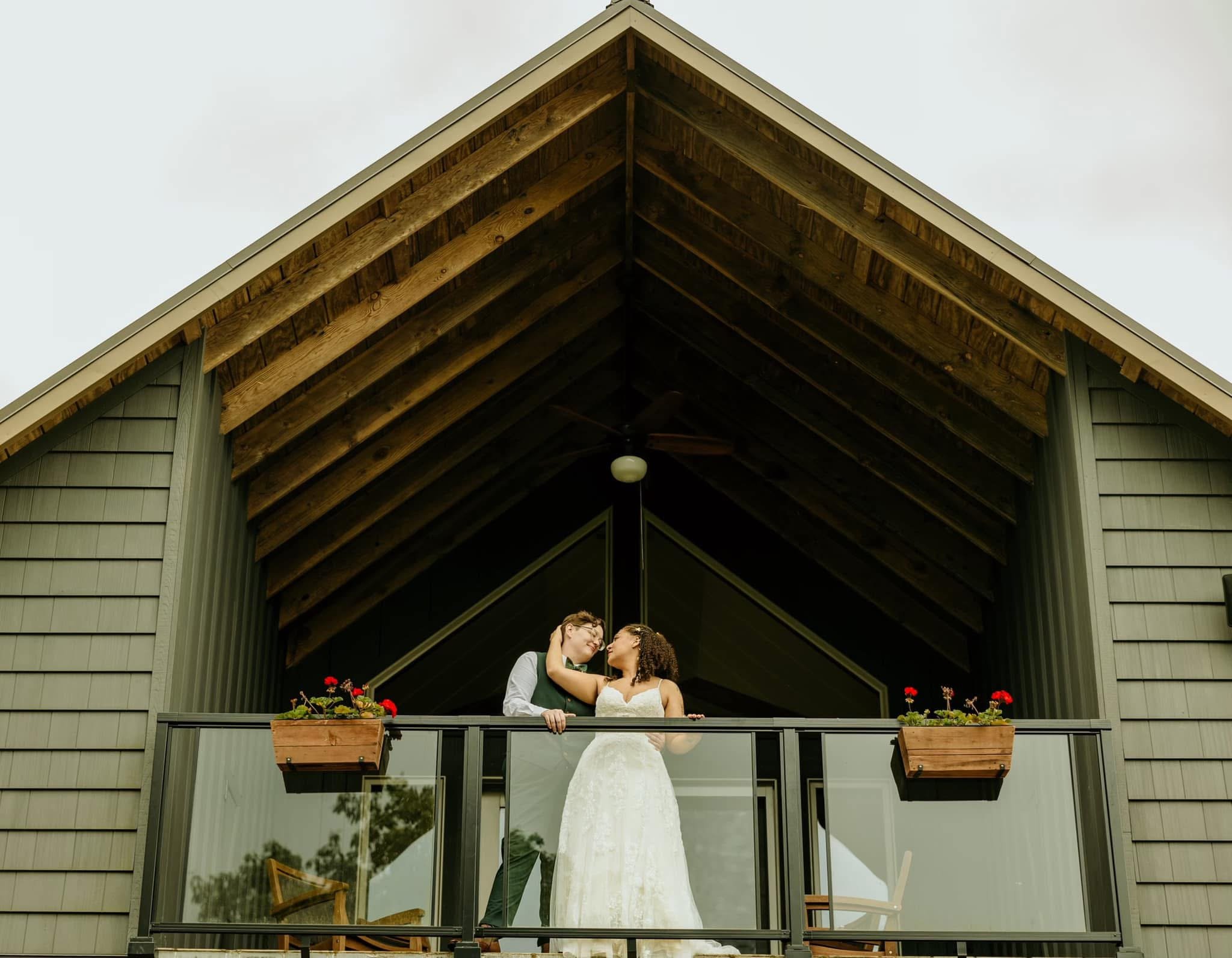 A couple, dressed in wedding attire, standing on a balcony of a modern house, sharing a romantic moment, with the house's roof and a cloudy sky in the background.