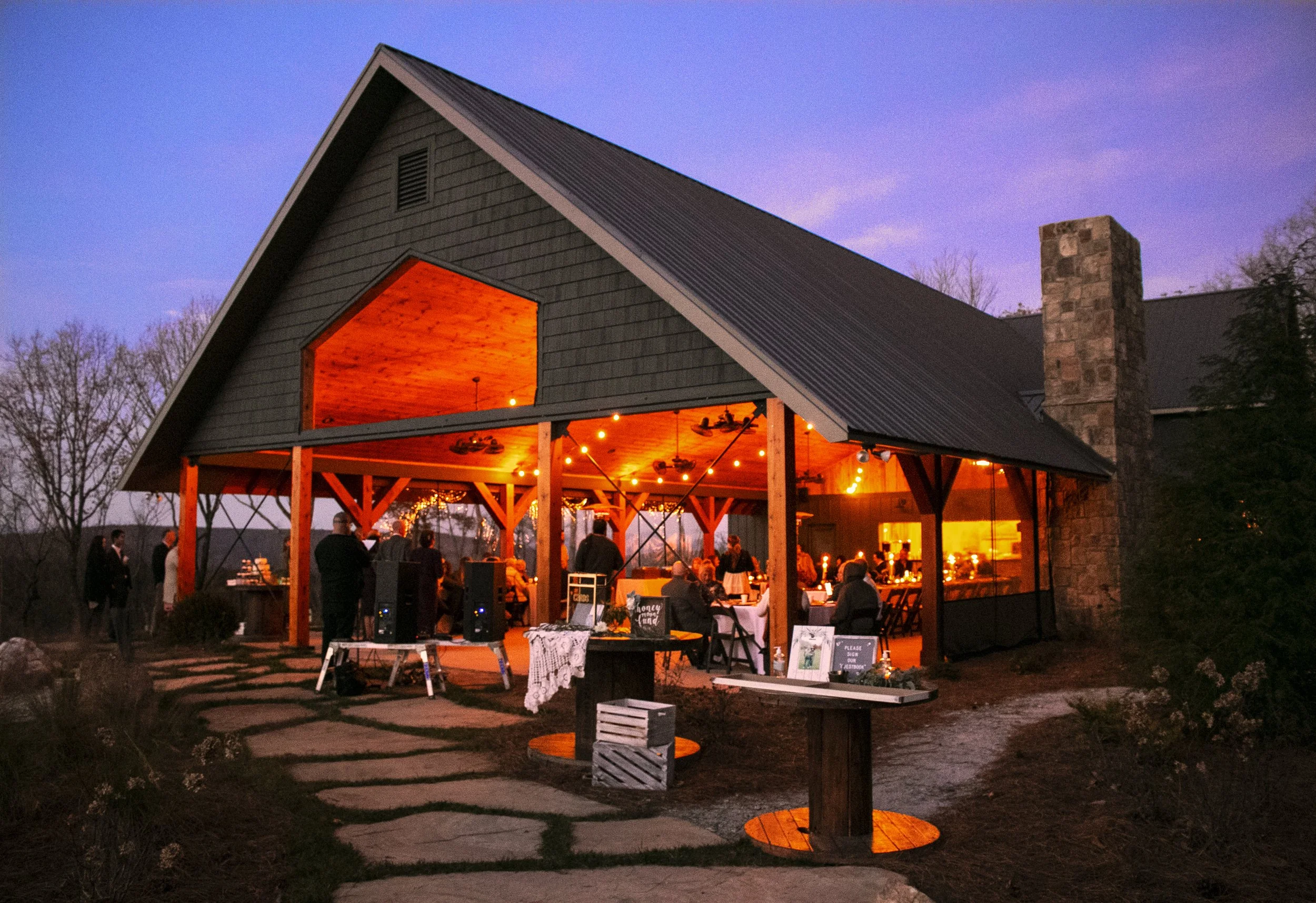 Outdoor barn wedding reception at dusk with warm lighting, tables, and guests.