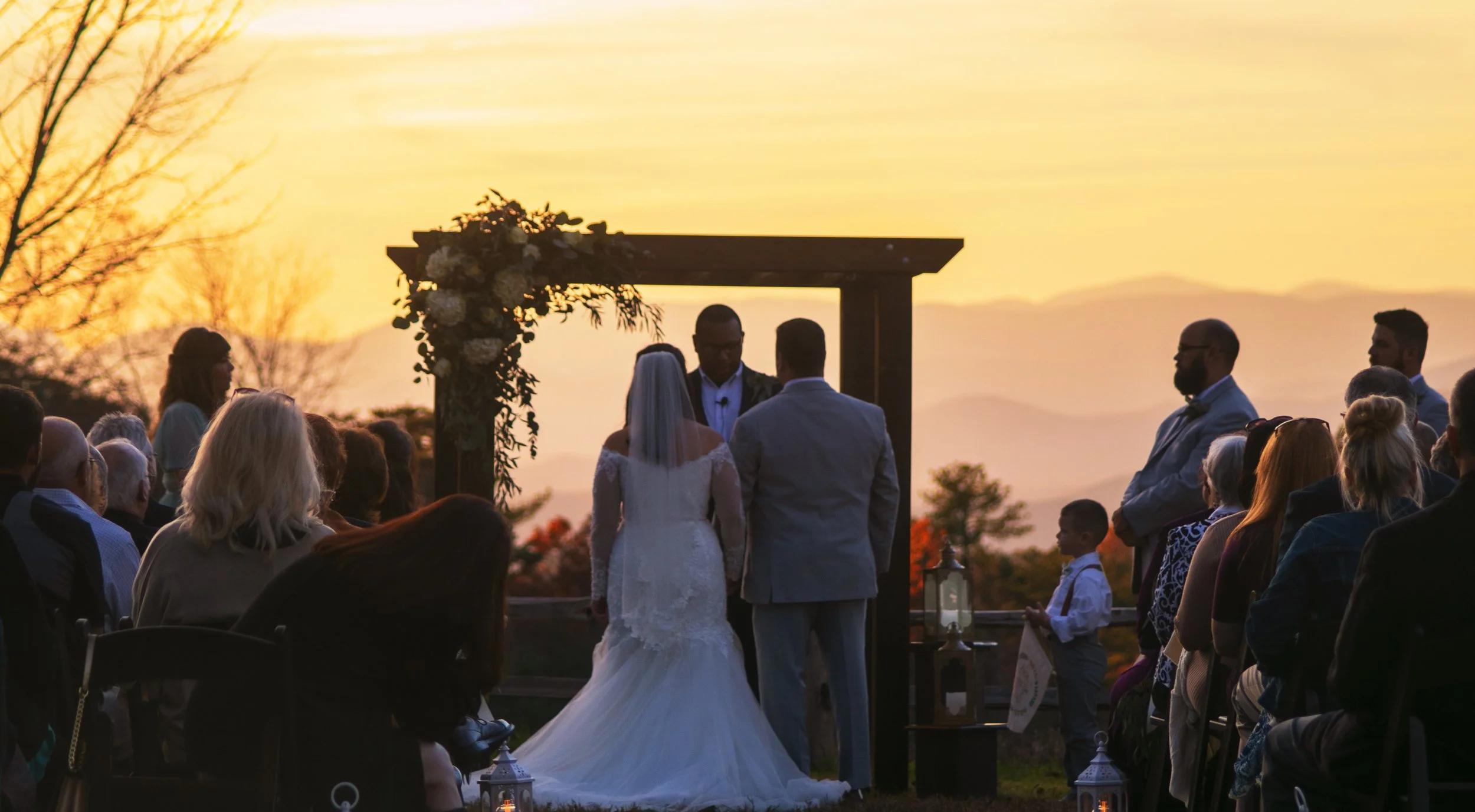 A couple getting married outdoors during sunset, standing under a wooden arch decorated with flowers, with guests seated on either side watching the ceremony.