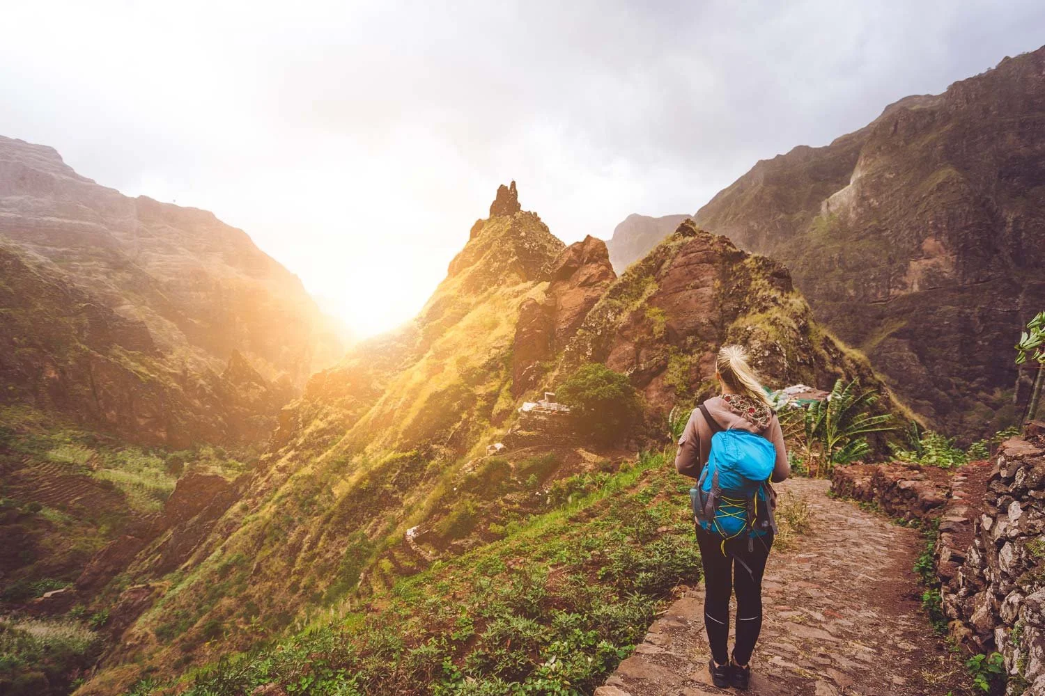 A woman with a blue backpack walking on a stone trail in a mountainous area with green vegetation, during sunrise or sunset.