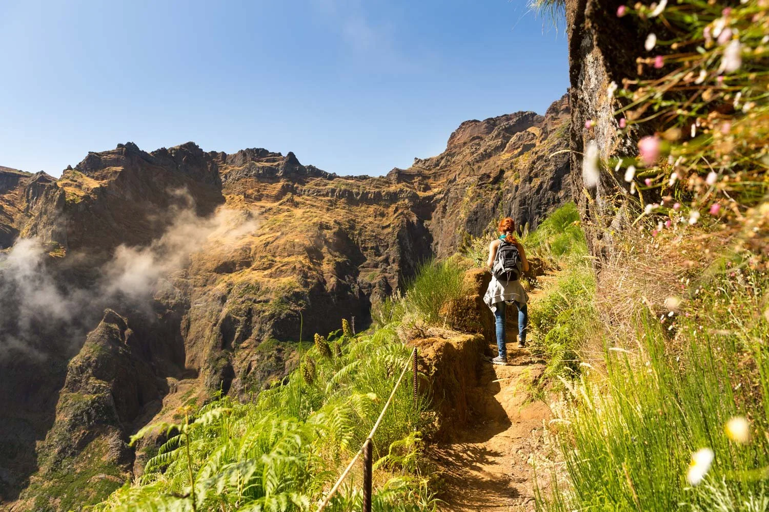 A woman with a backpack hiking on a narrow trail along a lush, green cliffside with rocky mountains in the background under a clear blue sky.