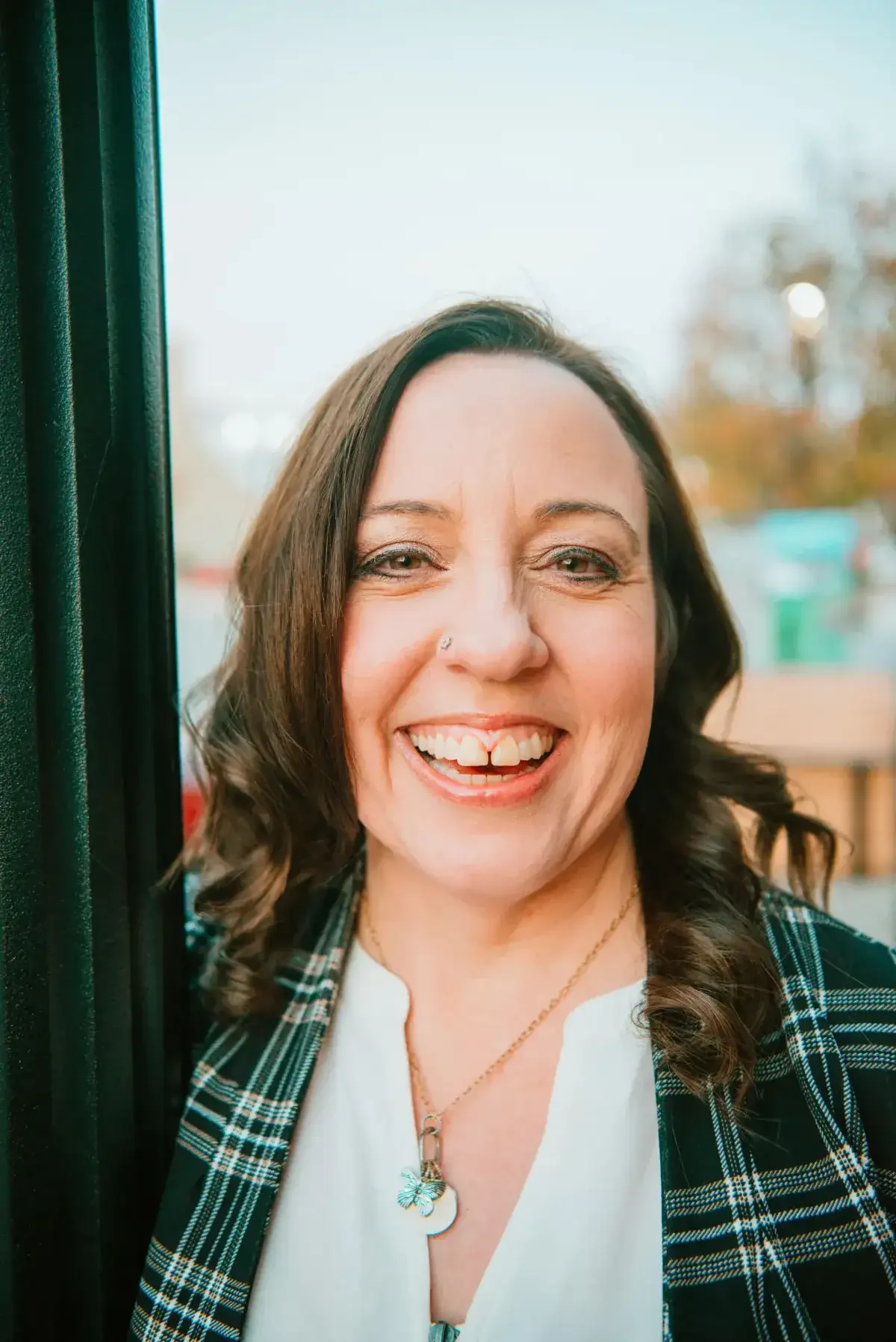 Close-up of a smiling woman with dark hair, wearing a white blouse, a plaid blazer, and a necklace with a butterfly and crescent moon pendant, standing outdoors near a window.