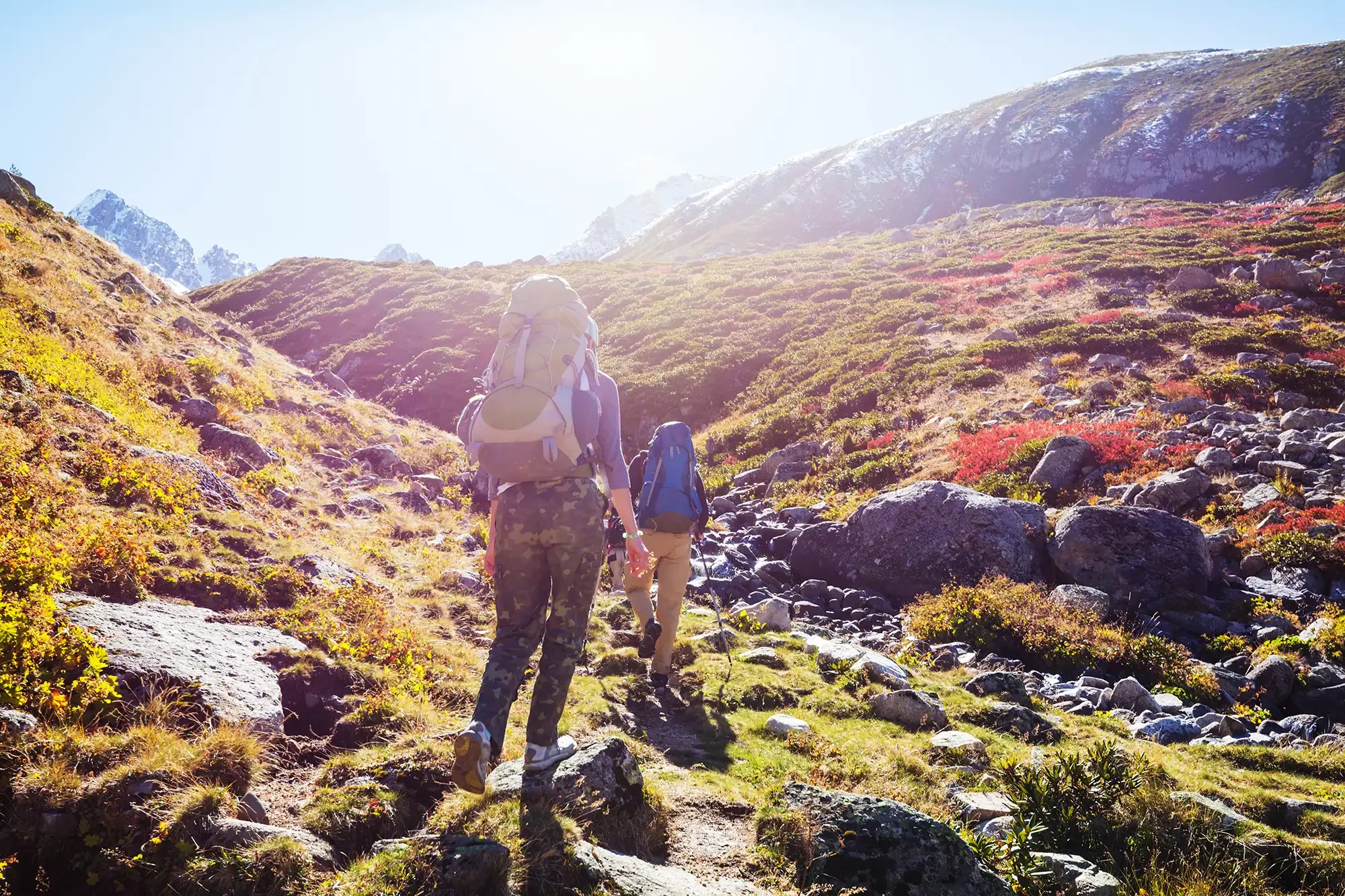 Two hikers with backpacks ascending a steep, rocky mountain trail surrounded by colorful vegetation and rocks, with snow-capped mountains in the background under a bright sky.