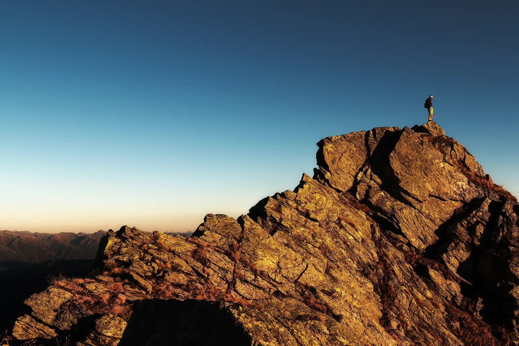 A hiker standing on the edge of a rocky mountain peak at sunset or sunrise, with the sky clear and distant mountain ranges in the background.